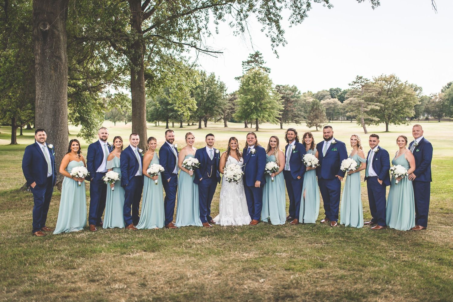 Wedding party in navy suits and light blue dresses posed together on grassy lawn.