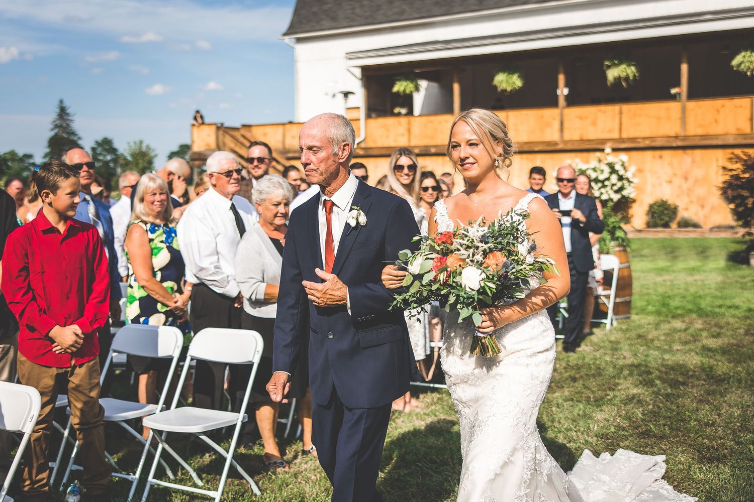 Wedding ceremony guests seated in white chairs on a lawn with rustic barn buildings in the background.