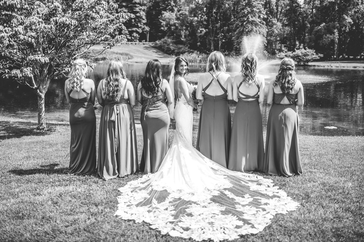 Wedding party gathered on grass with lake and fountain backdrop for formal group photos.