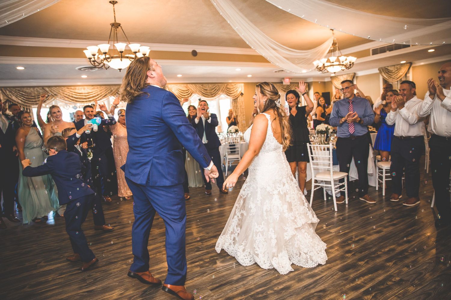 Wedding couple shares their first dance in an elegant ballroom with chandeliers and guests watching.