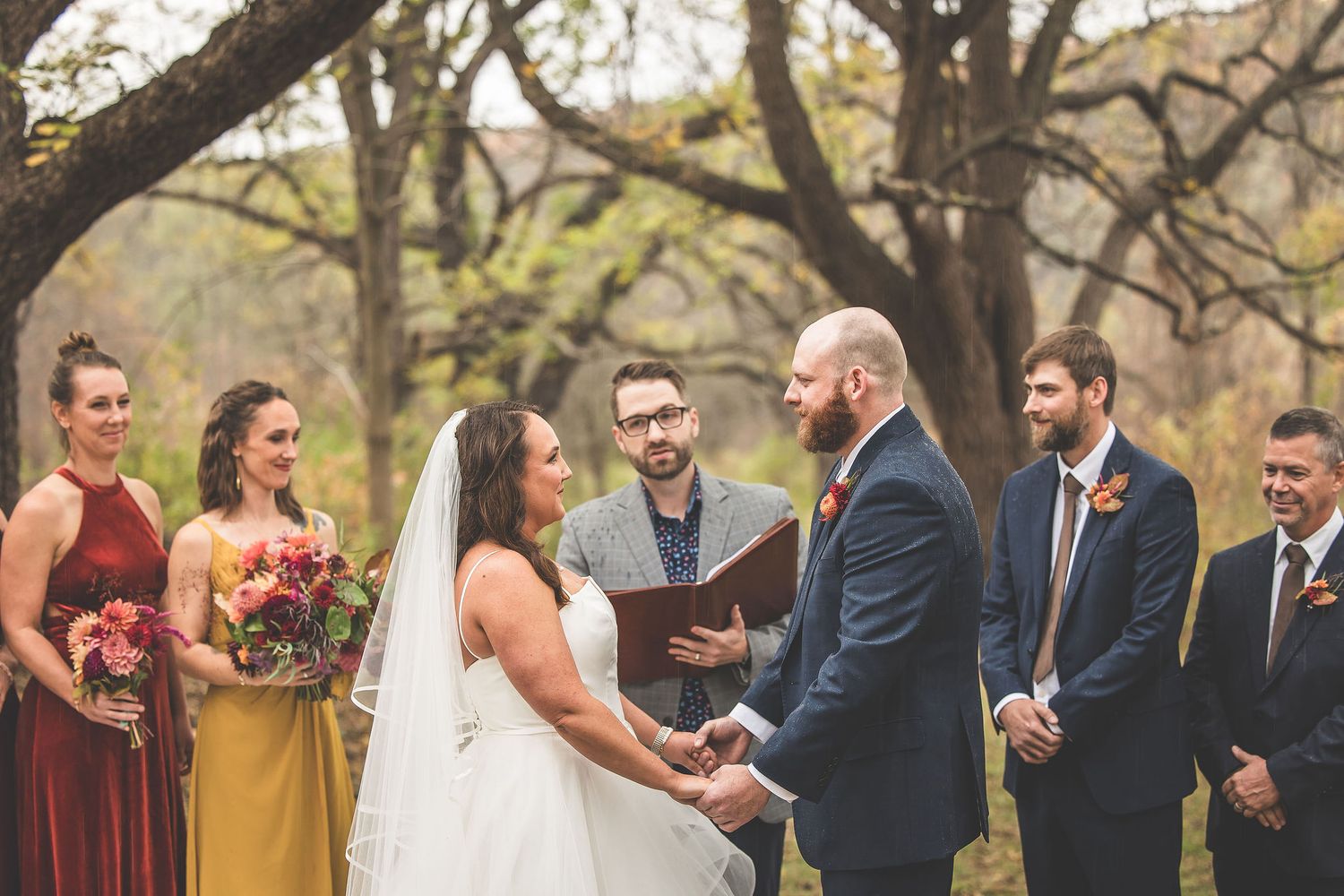 An outdoor wedding ceremony takes place under autumn trees with burgundy and gold bridesmaids dresses.