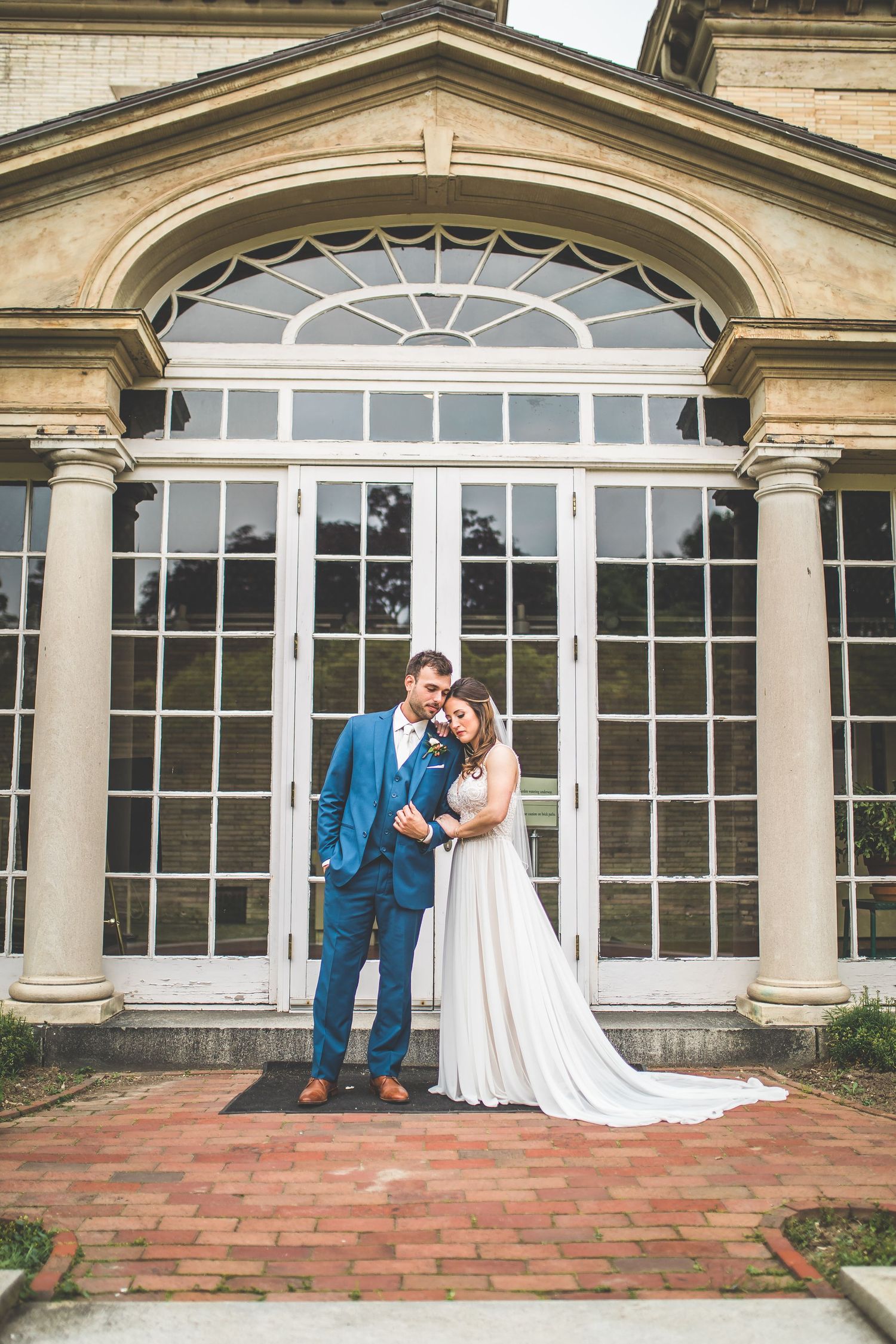 A couple stands before an elegant arched doorway with columns and brick path on their wedding day.