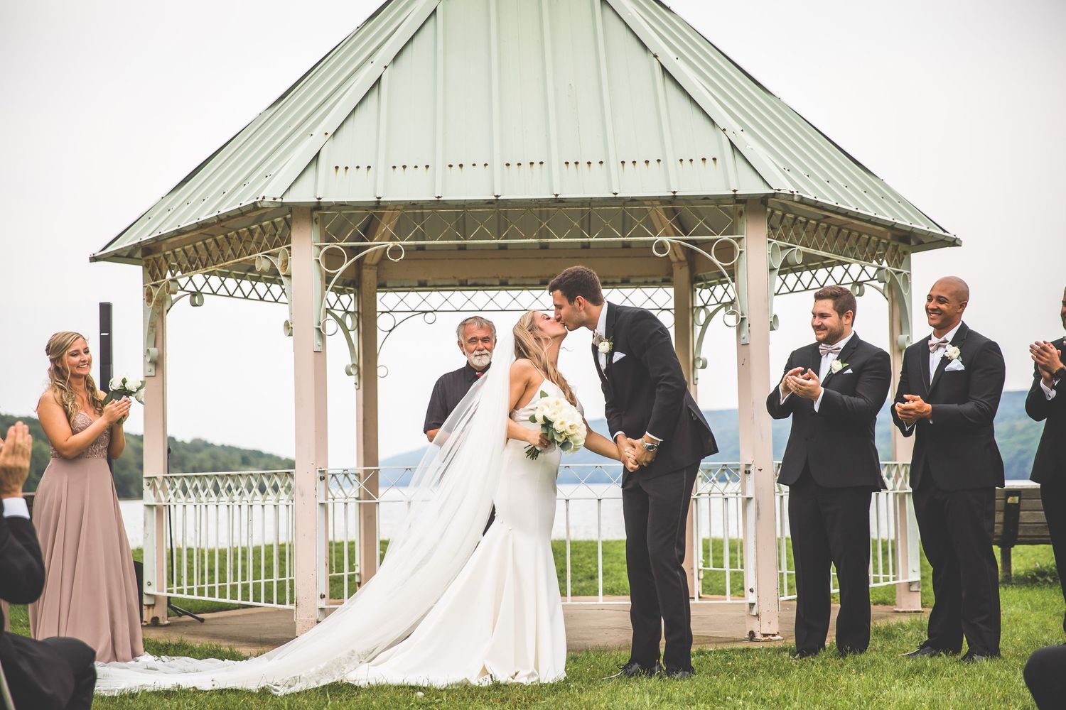 Scenic lakeside wedding ceremony with white gazebo and formal gathering of guests in formal attire.