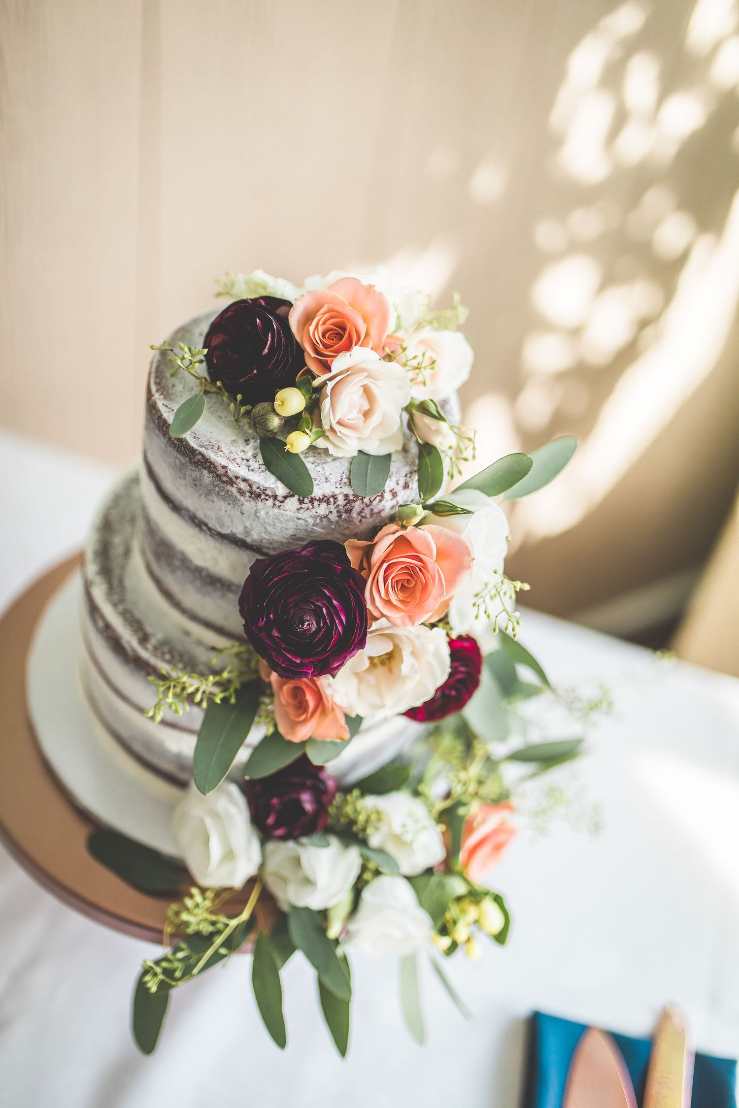 Semi-naked wedding cake decorated with peach roses and deep burgundy flowers.