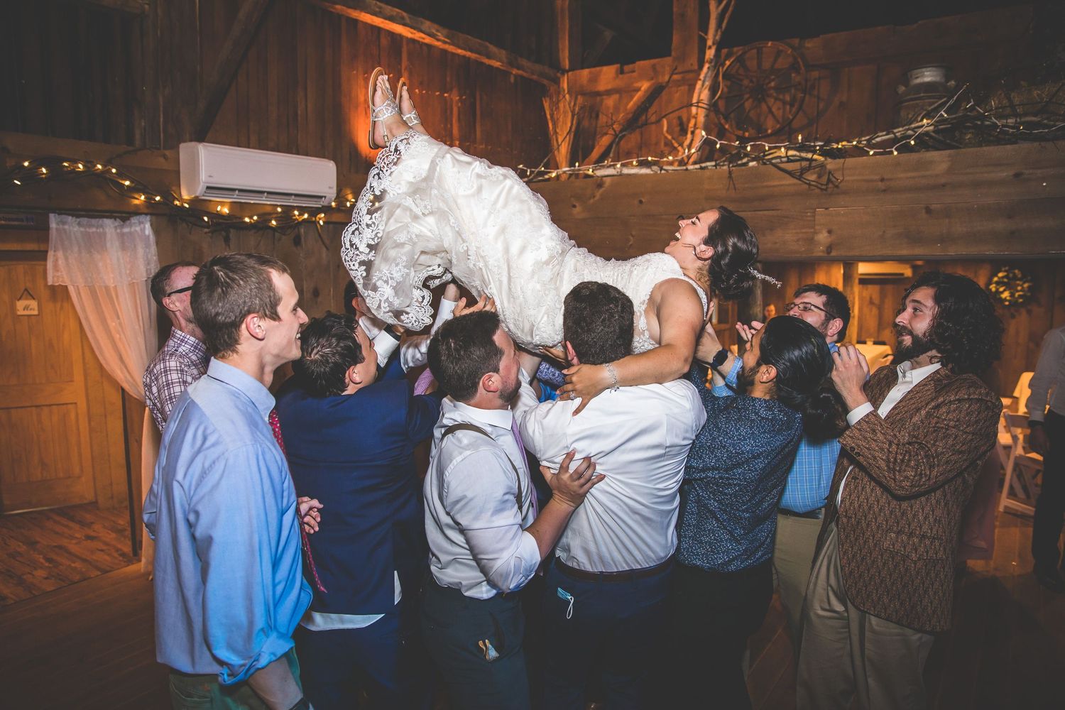 Panoramic view of wedding guests dancing in a barn decorated with string lights.