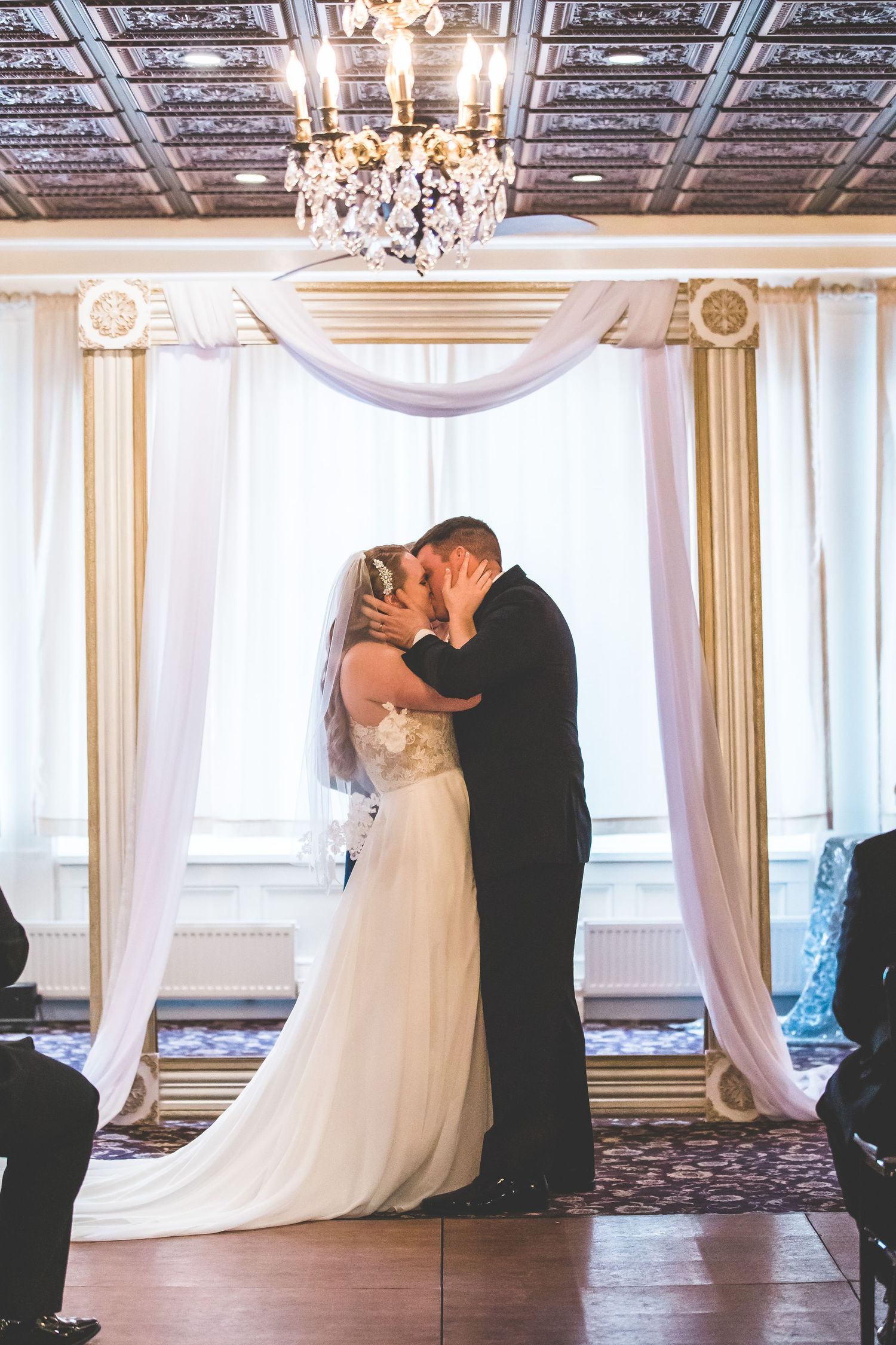 Romantic first kiss during wedding ceremony under ornate ceiling and crystal chandelier.