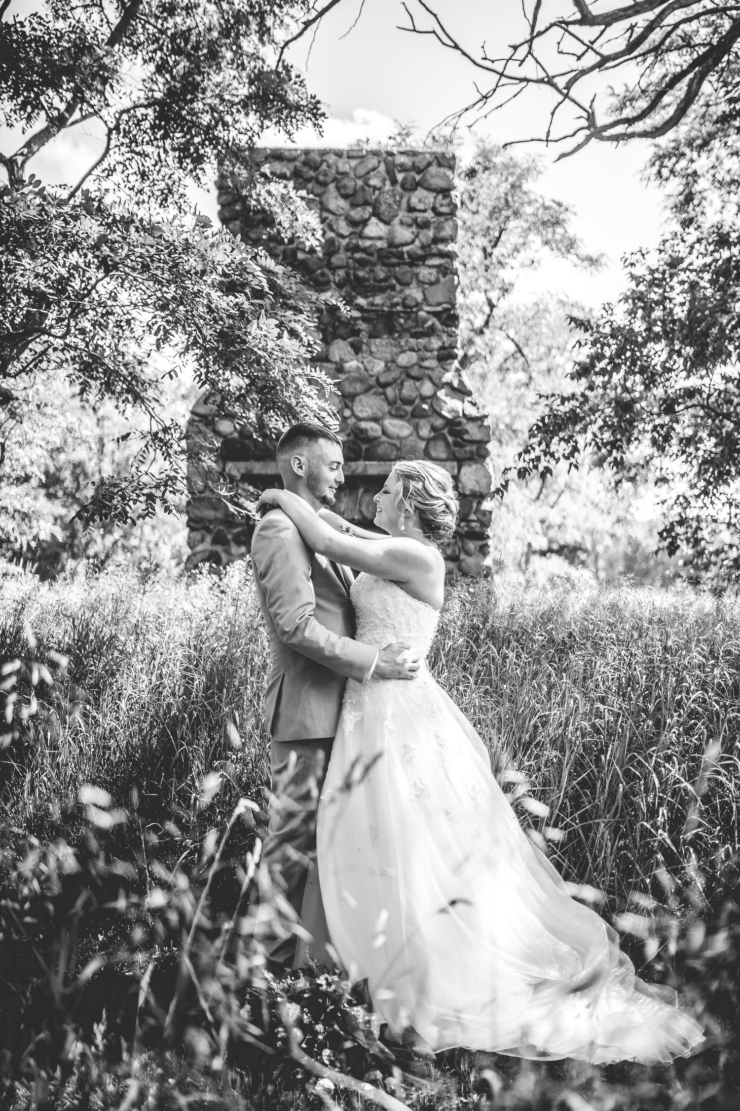 Black and white wedding portrait with a couple embracing near a stone wall in a meadow.
