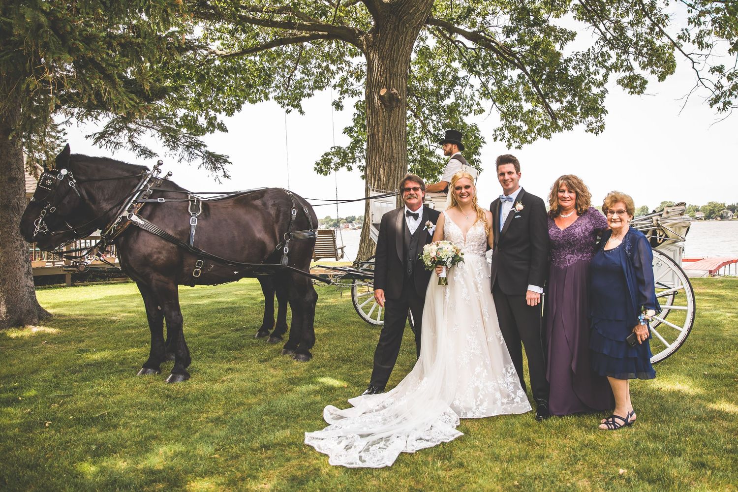 Wedding party poses outdoors under large trees near a waterfront setting.
