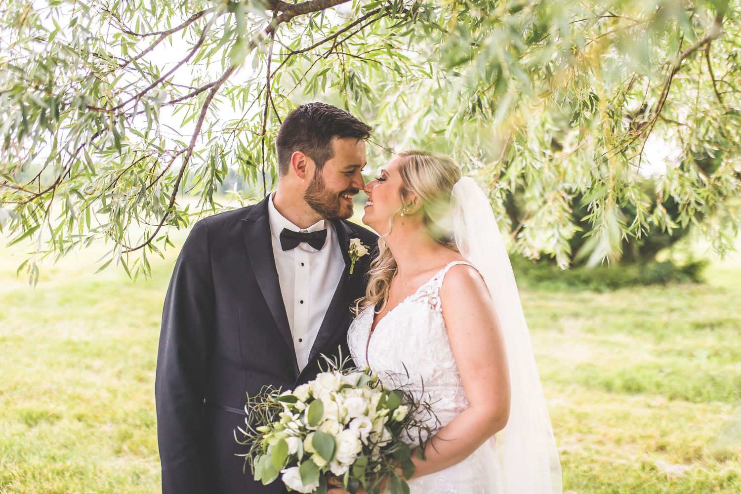 Romantic wedding portrait of couple with white bouquet beneath drooping willow branches.