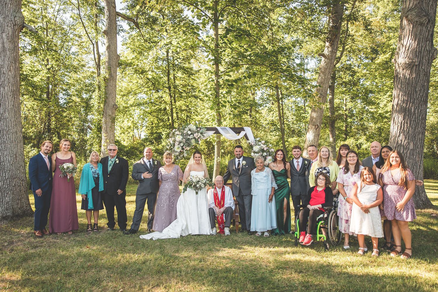 Large group wedding photo taken outdoors with trees and natural lighting.