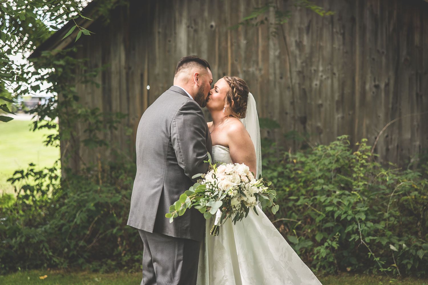 Intimate wedding moment captured beside weathered barn with trailing white dress.