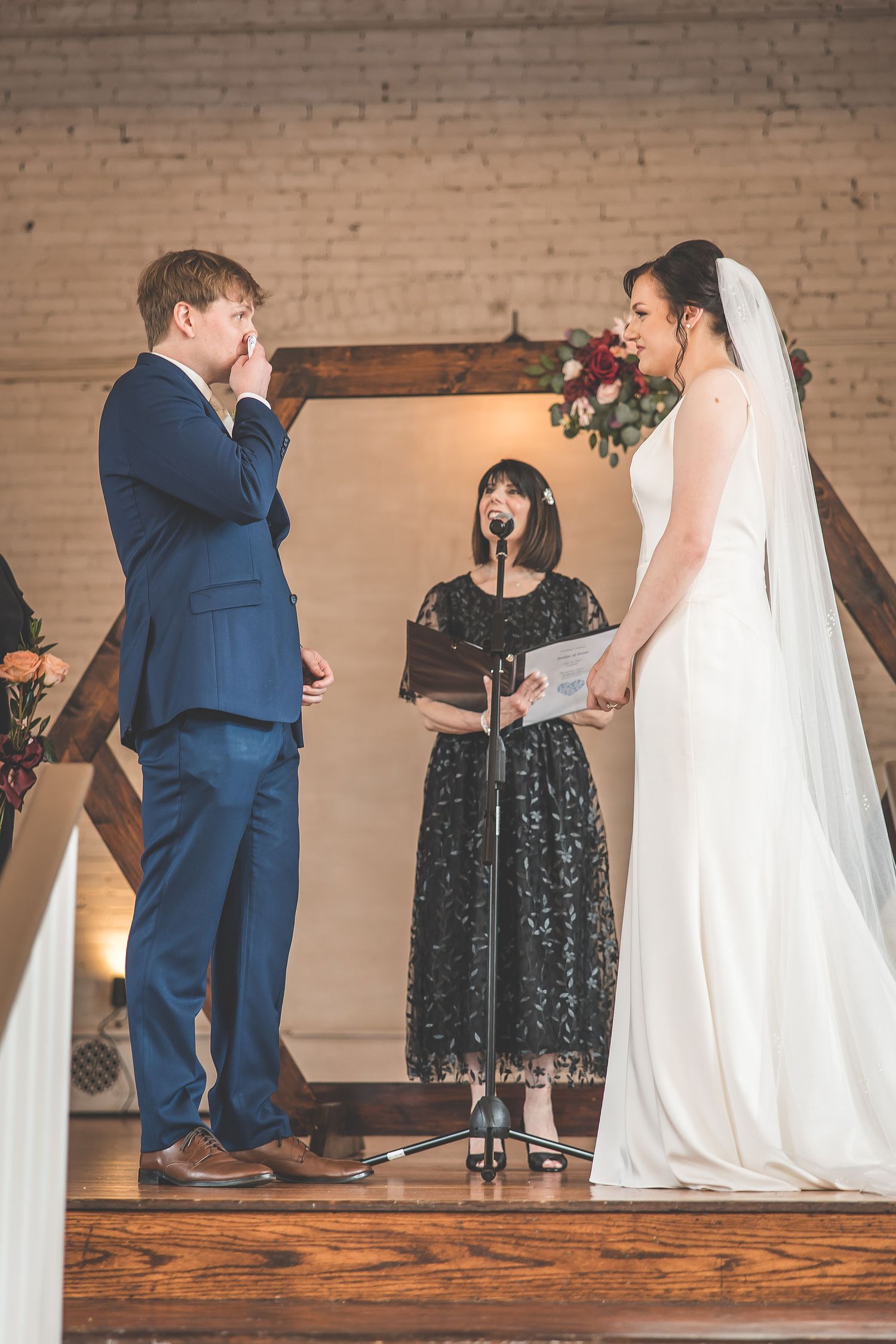 Wedding party lined up on wooden stage during indoor ceremony with brick wall background.