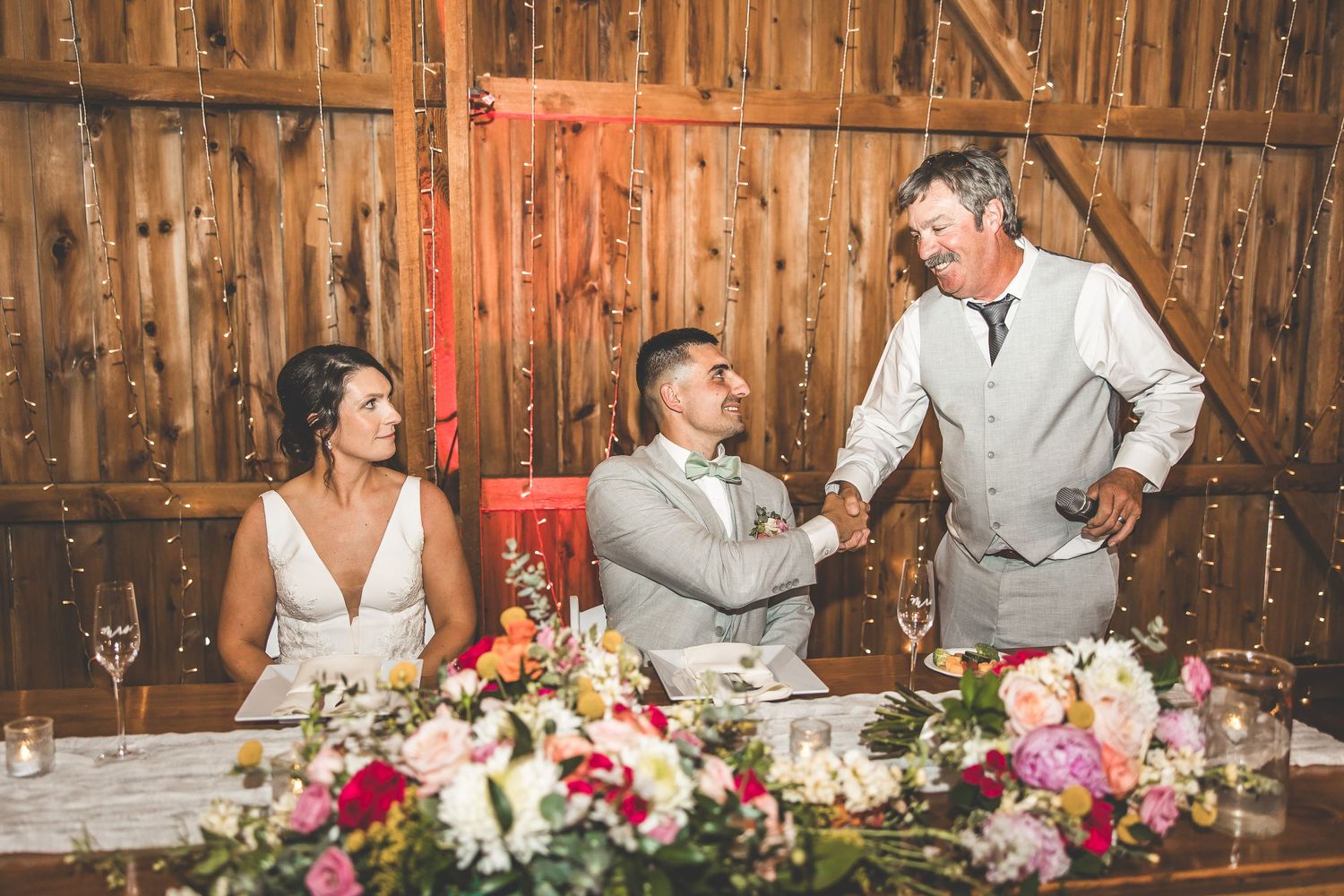 Wedding reception dinner scene with guests and floral centerpieces in a rustic barn setting.