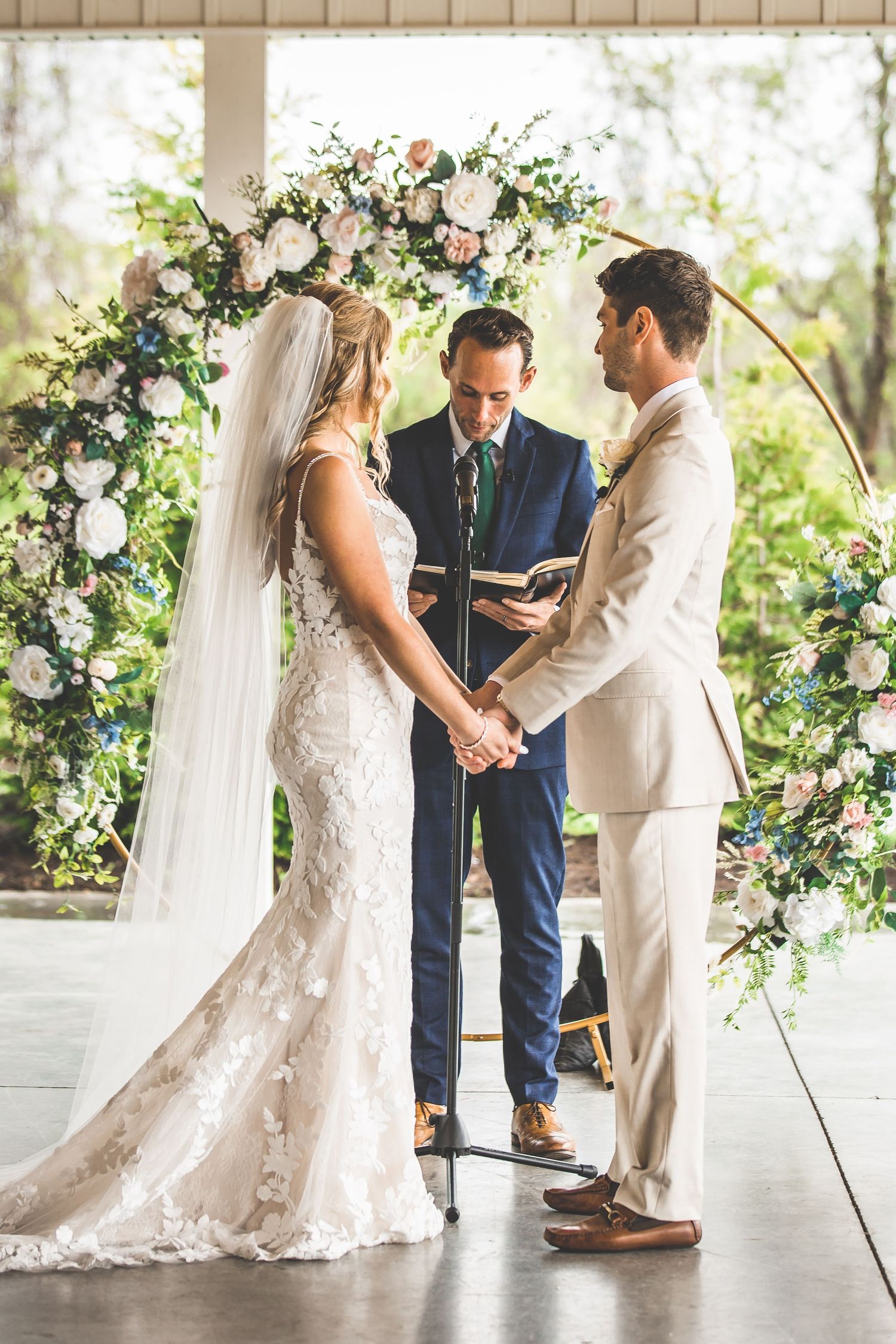 Wedding ceremony takes place under floral arch decorations in bright indoor venue.