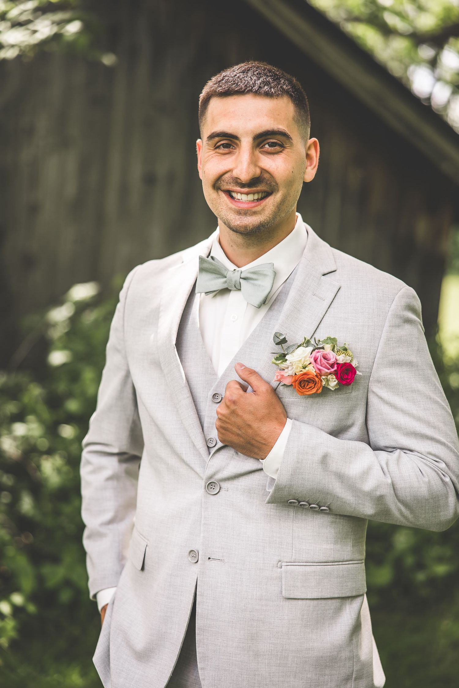 Groom in light grey suit adjusts his tie while smiling outdoors.