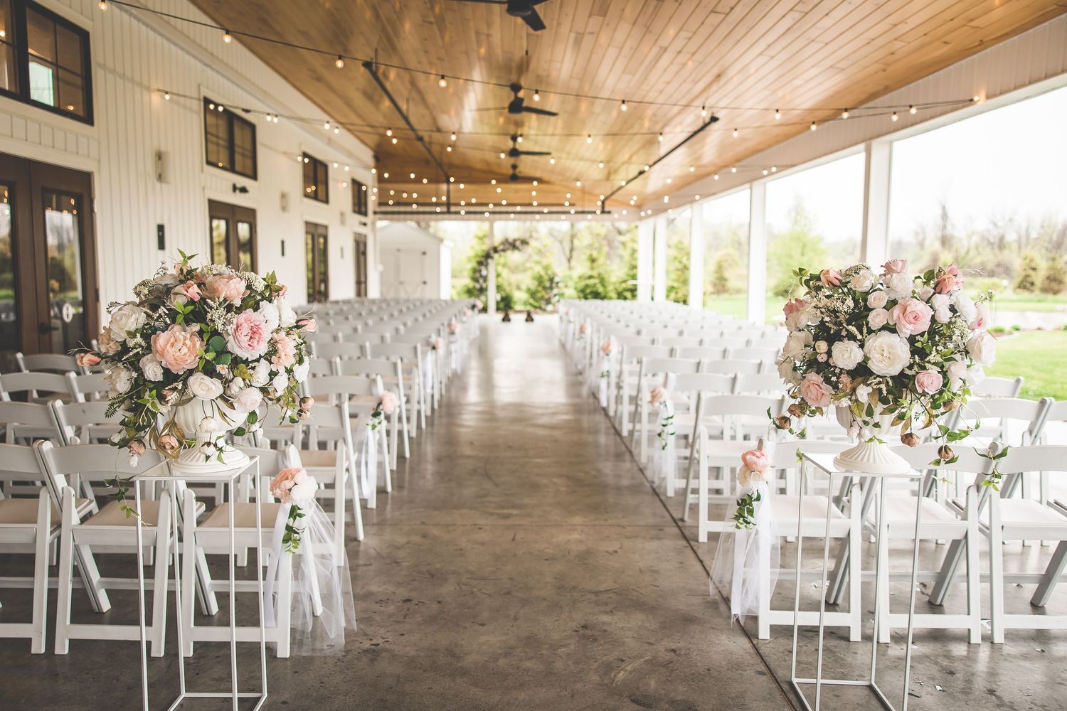 An elegant wedding ceremony setup with white chairs and pink floral arrangements in a covered outdoor venue.