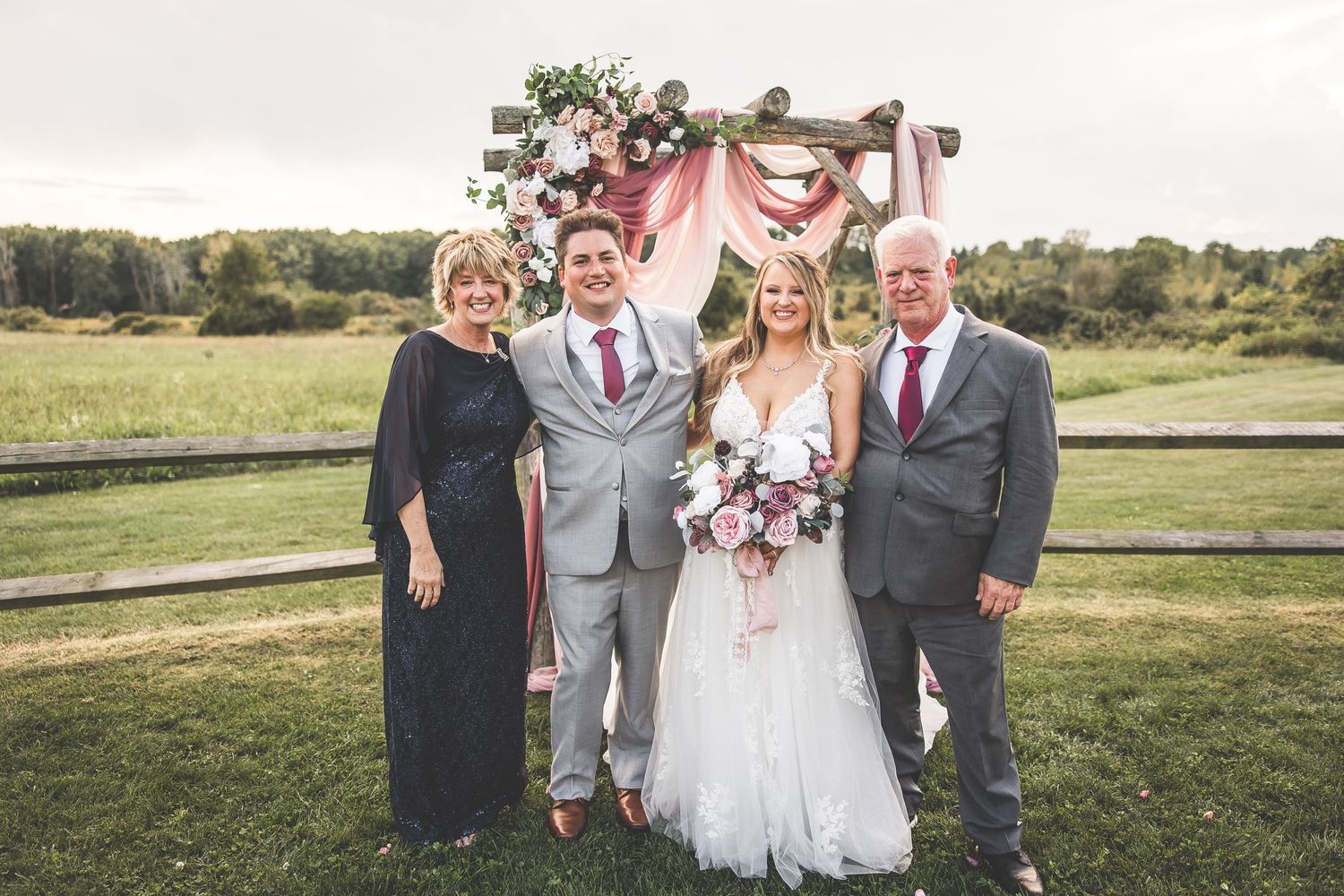 Wedding group photo with bridal party wearing pink dresses and gray suits in a scenic field.