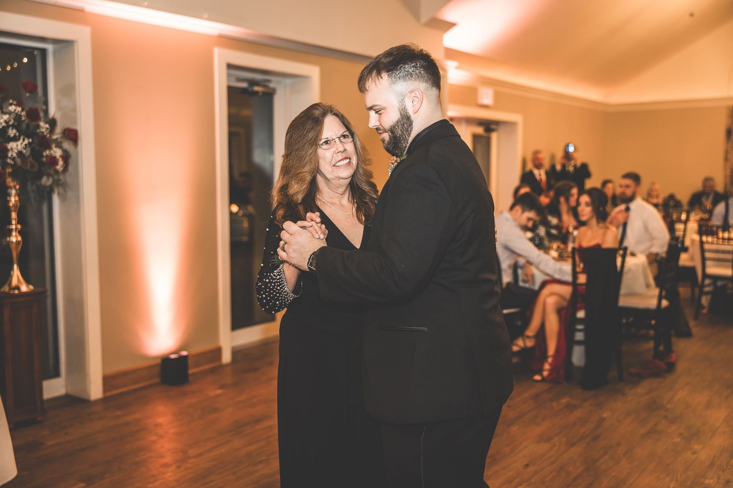 Wedding celebration in a warmly lit reception hall with guests dancing on hardwood floors.