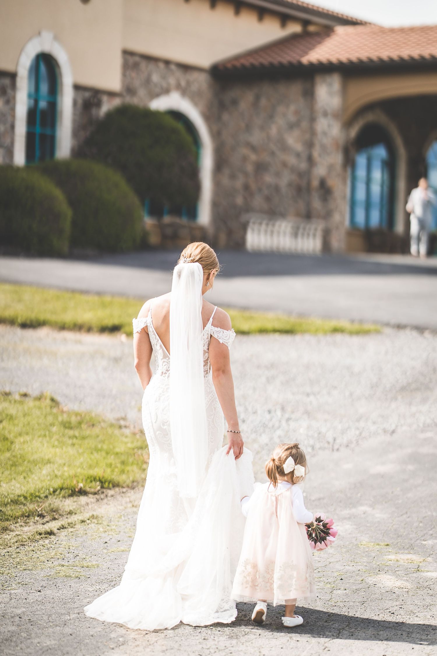 Bride and flower girl walking together on stone path outside Mediterranean-style building.