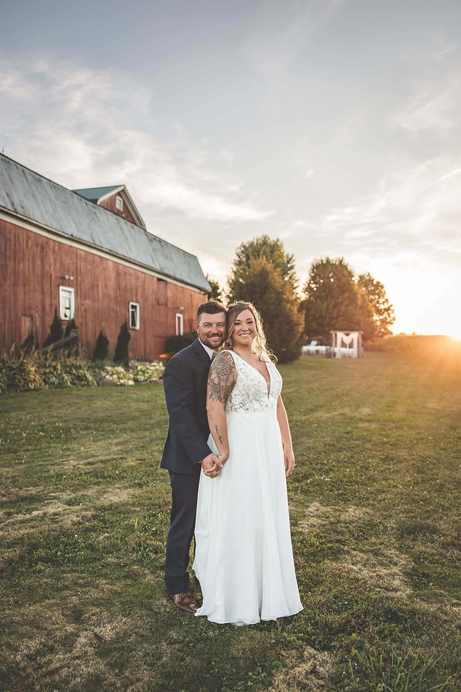Newlyweds pose in front of rustic red barn during golden hour sunset.