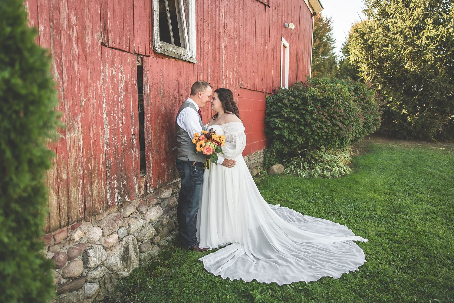 Wedding photos captured against red barn with flowing white dress train.