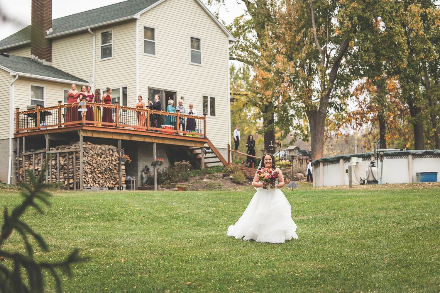 A wedding couple poses outdoors on a grassy hill with white buildings and fall trees in the background.