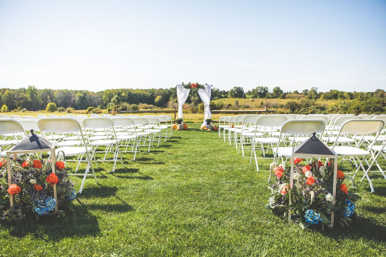 Outdoor wedding ceremony setup with white chairs and floral arrangements on a grassy field.