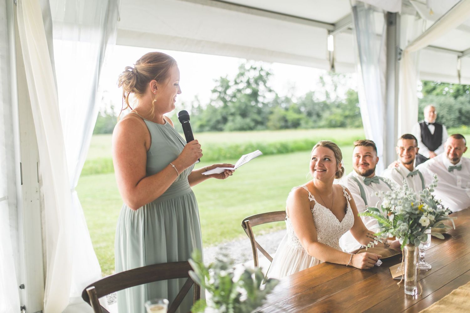 A wedding speech being delivered at a rustic outdoor reception under a white tent.