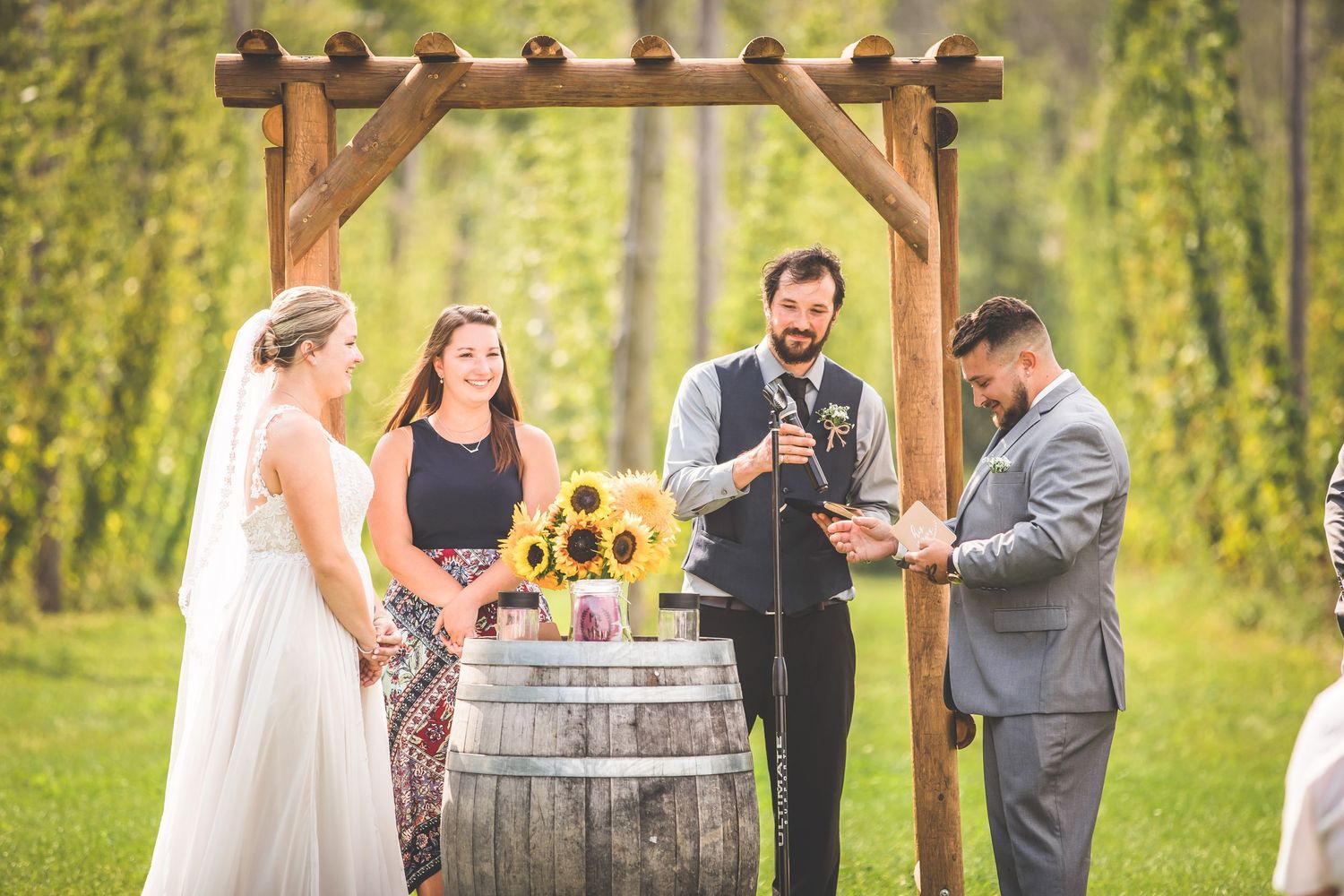 Wedding party gathered around wooden barrel altar during outdoor ceremony in grassy field.