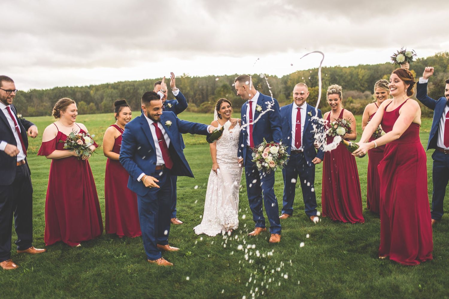 Wedding party stands in line on green grass wearing burgundy dresses and navy suits.