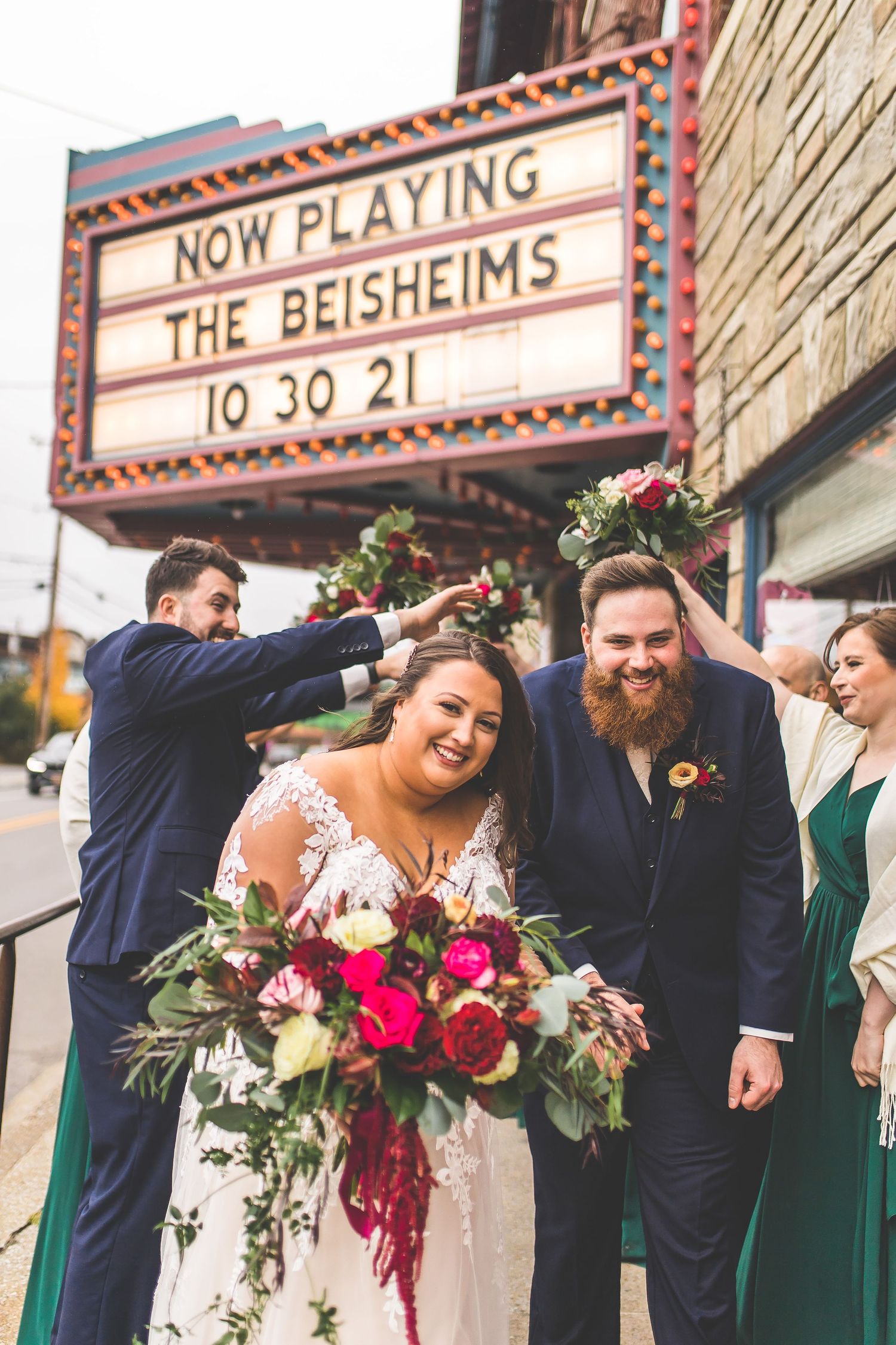 Wedding party in navy and emerald green attire strike playful poses together in front of a theater marquee.