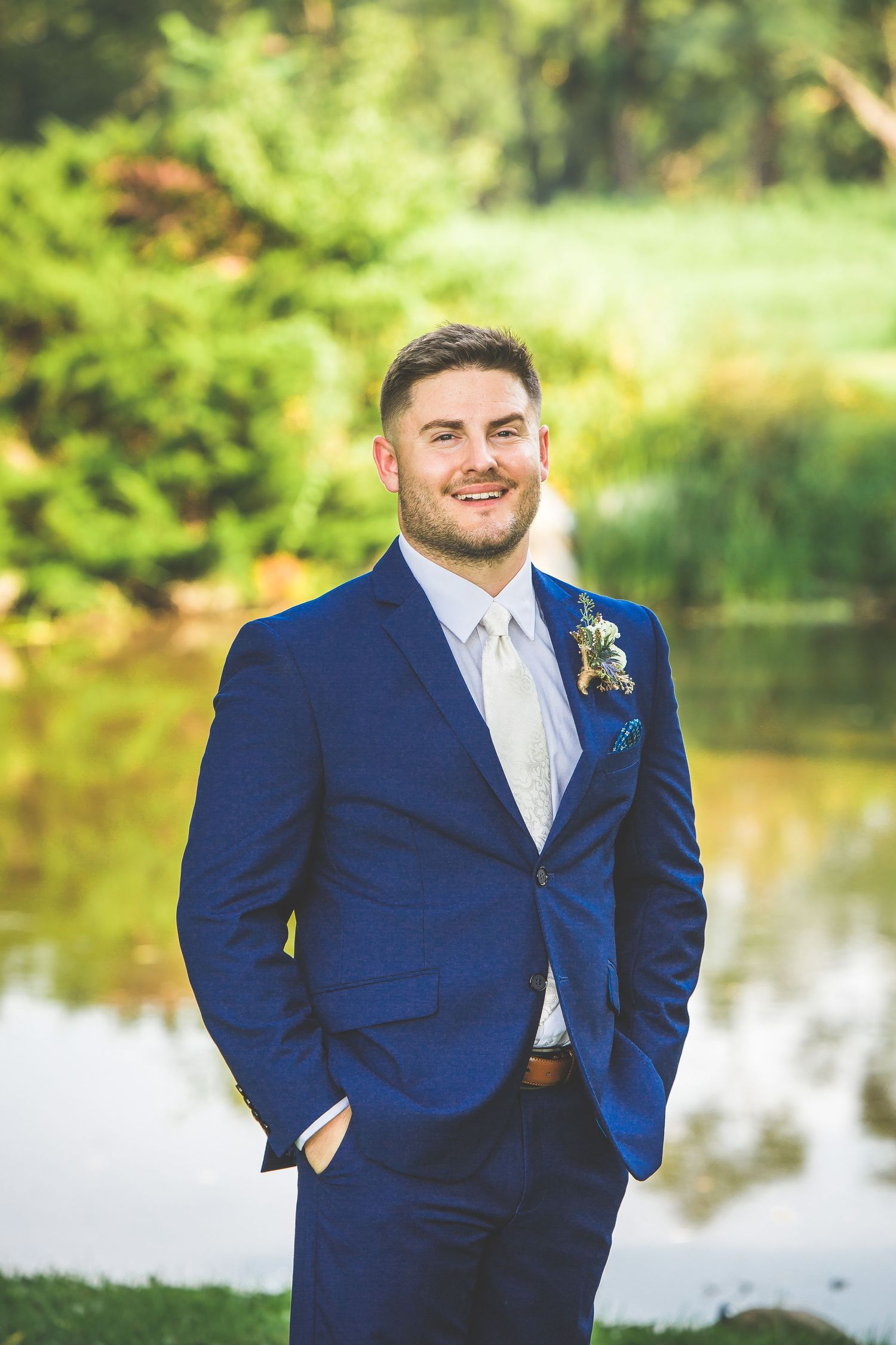 Groomsmen in navy blue suits pose near a reflective pond in a garden setting.