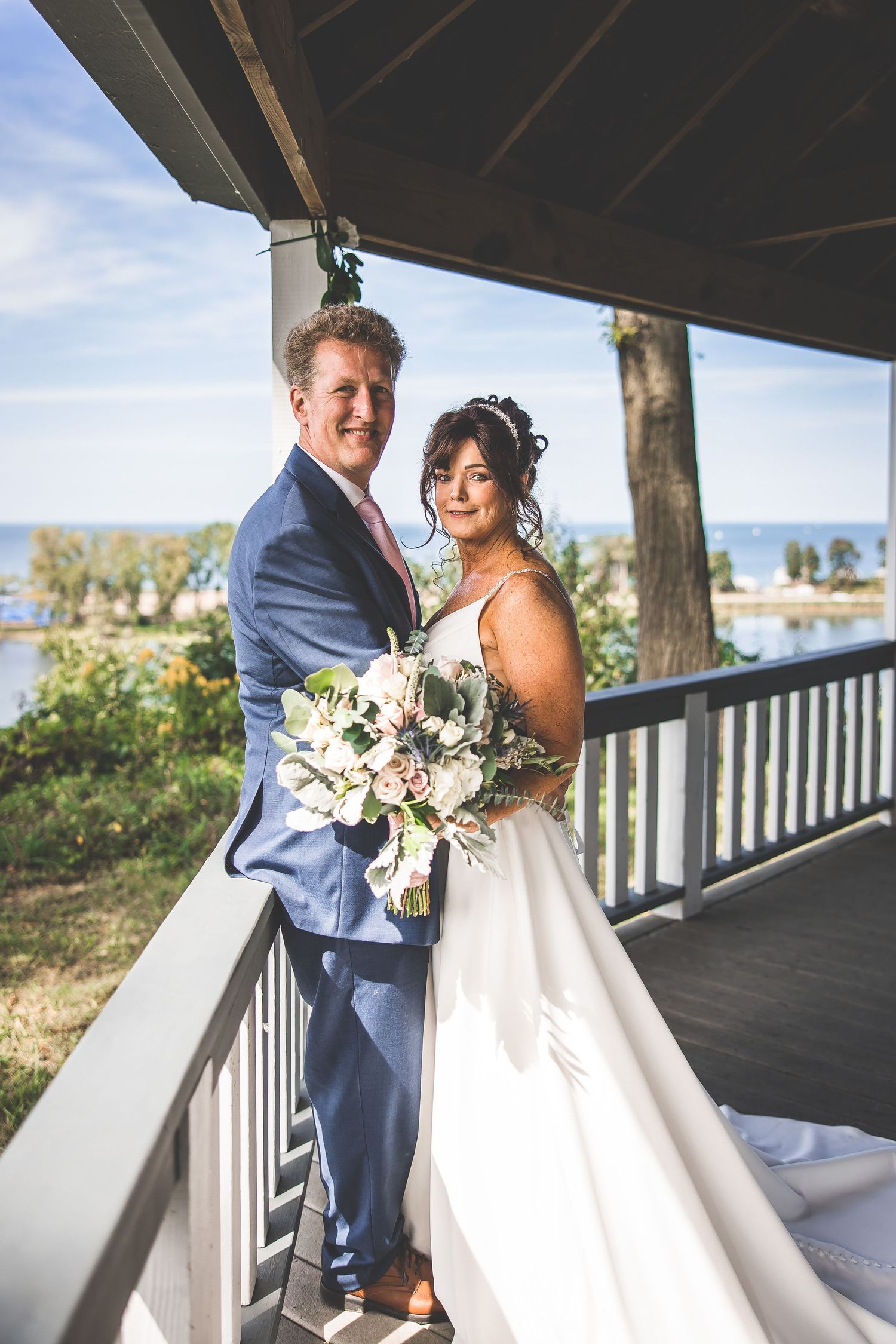 A couple embraces while holding a floral bouquet on a wooden deck overlooking the water.