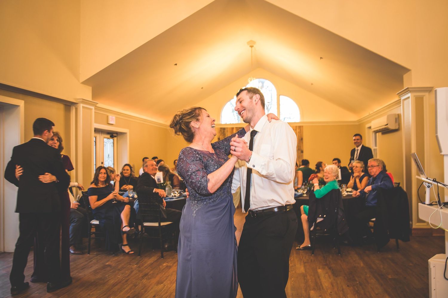 A couple shares a dance at a wedding reception in a warmly lit venue with vaulted ceilings.