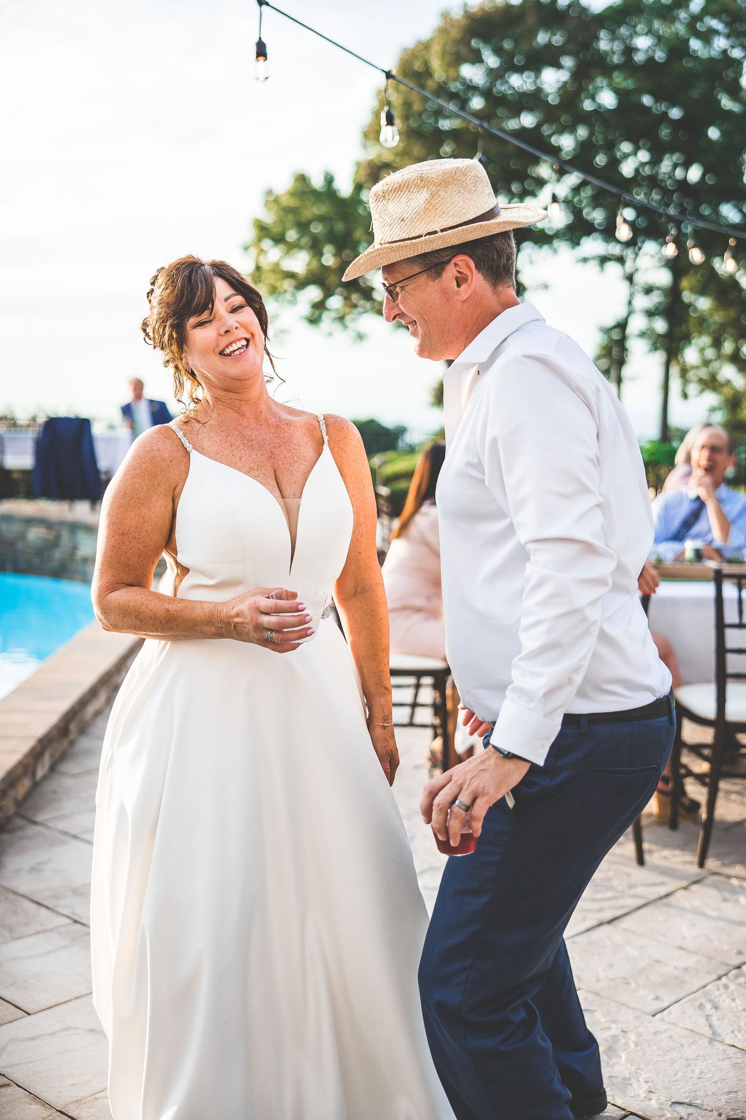 A couple dances and laughs together during their outdoor wedding reception by a pool.