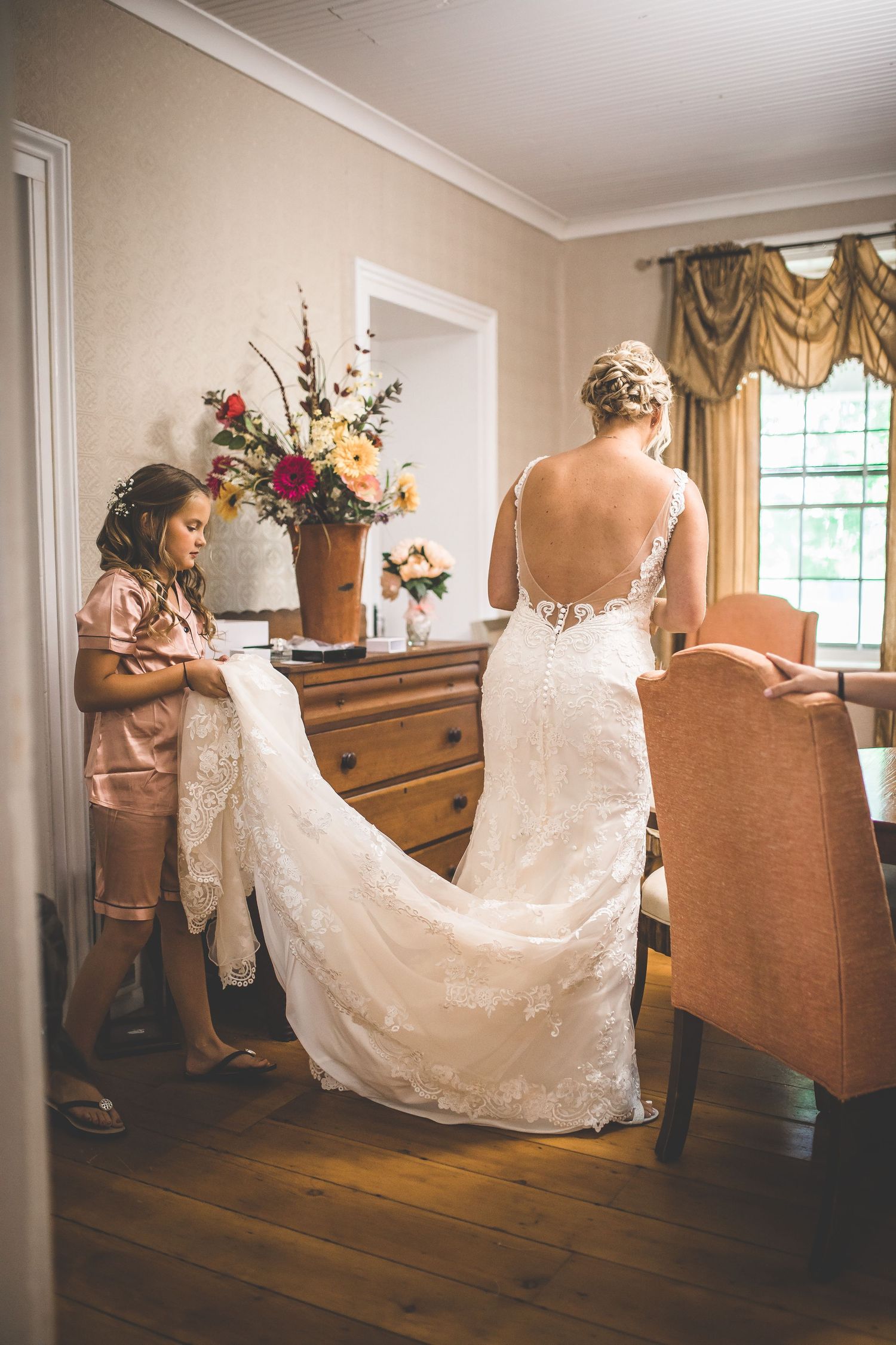 A bride in a backless lace wedding dress stands in an elegant room while her train is being arranged.