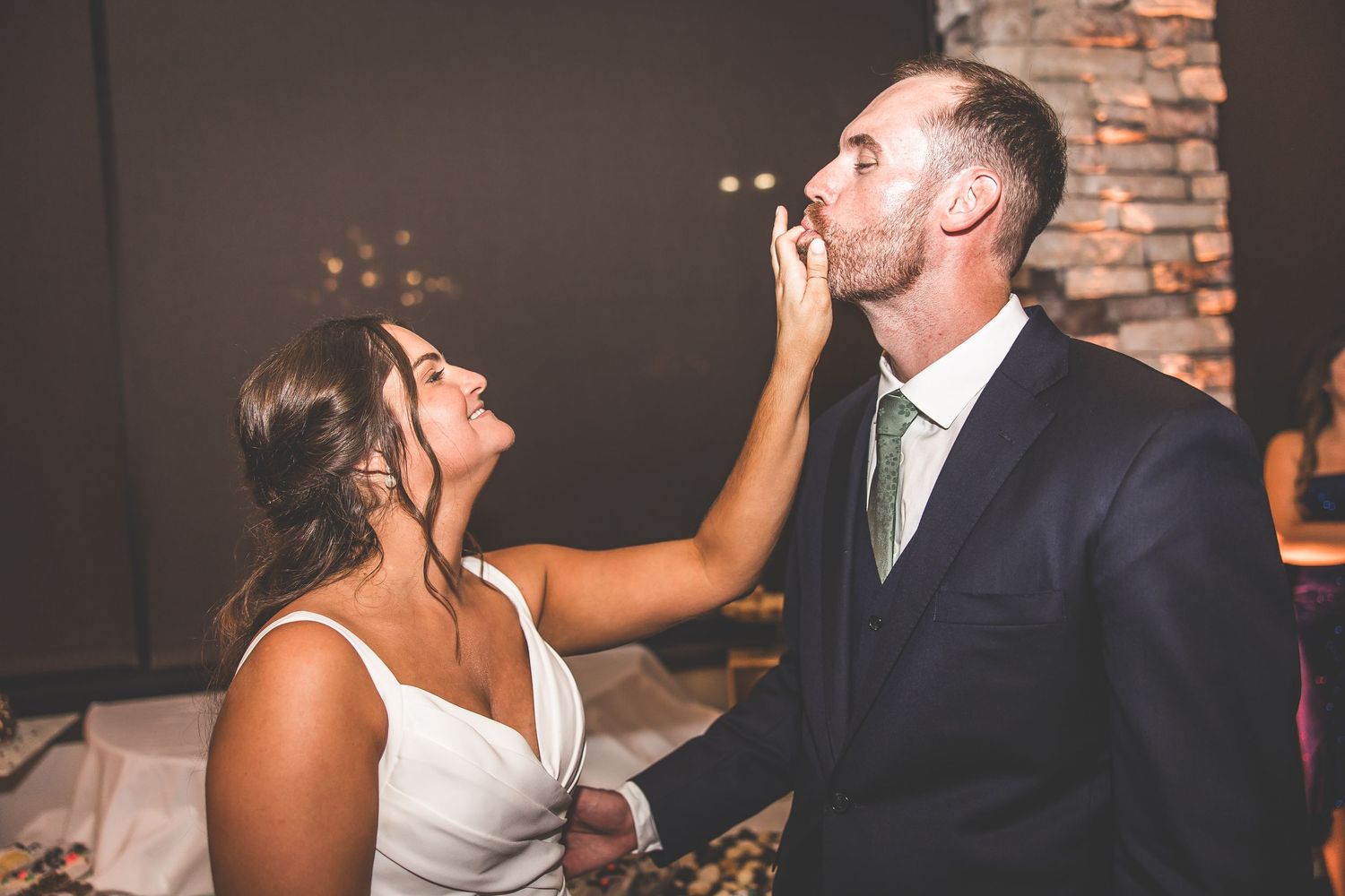 Playful cake smashing moment between newlyweds at indoor reception with brick wall backdrop.