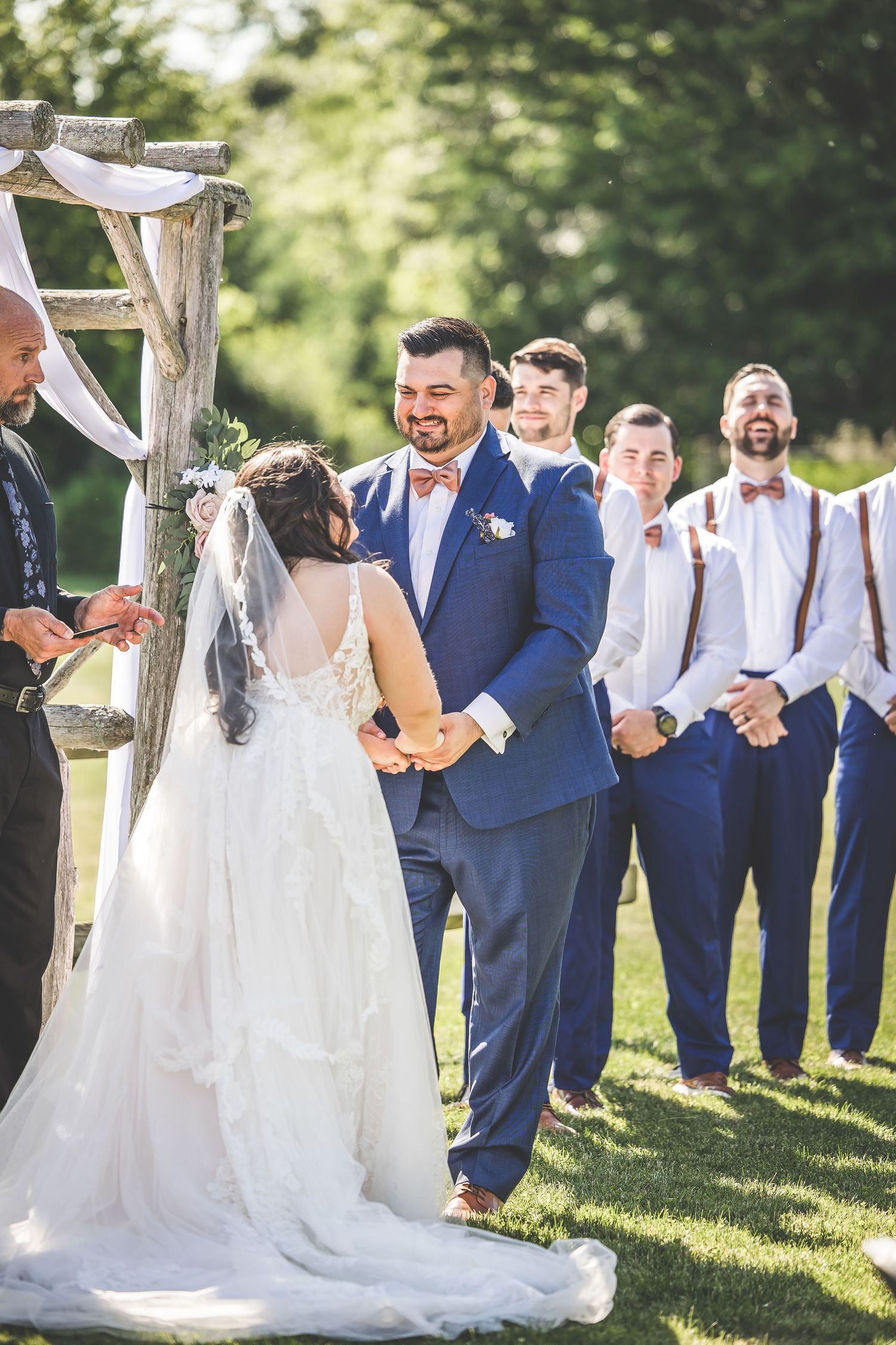 Wedding party in navy suits and white dresses poses outdoors under a rustic wooden arch with white fabric draping.