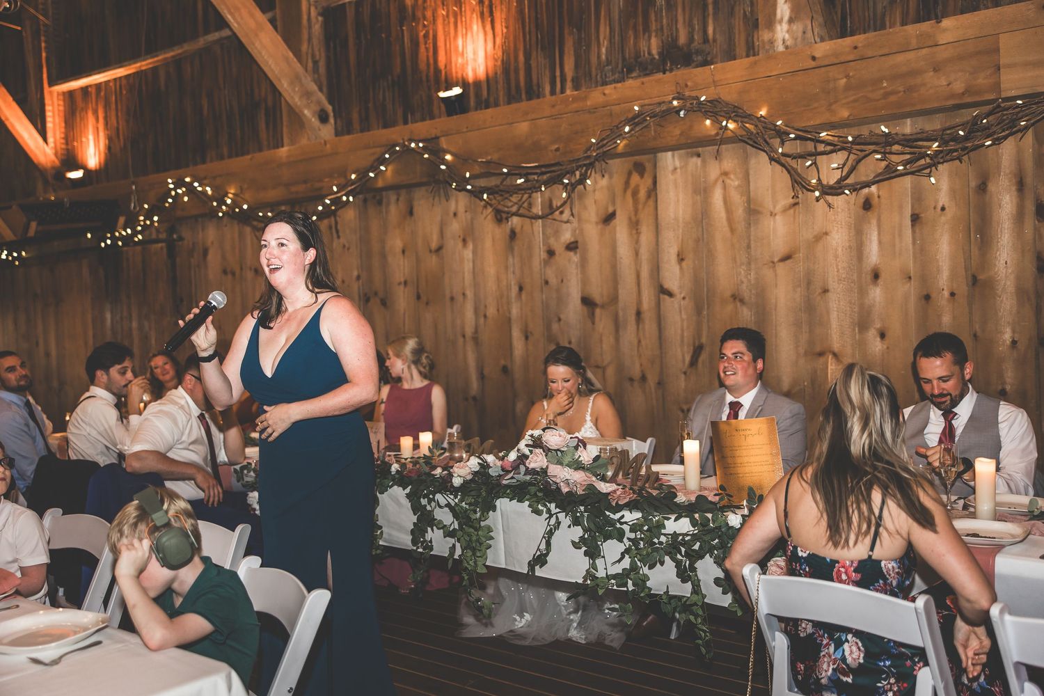 Guest giving speech at wedding reception in rustic barn decorated with string lights and floral arrangements.