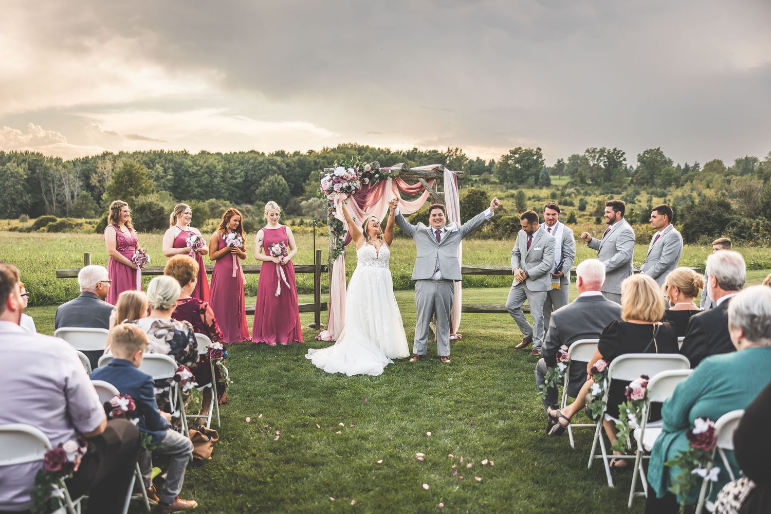 Wedding guests celebrate outdoors during a summer ceremony with white chairs set up on a grassy field.