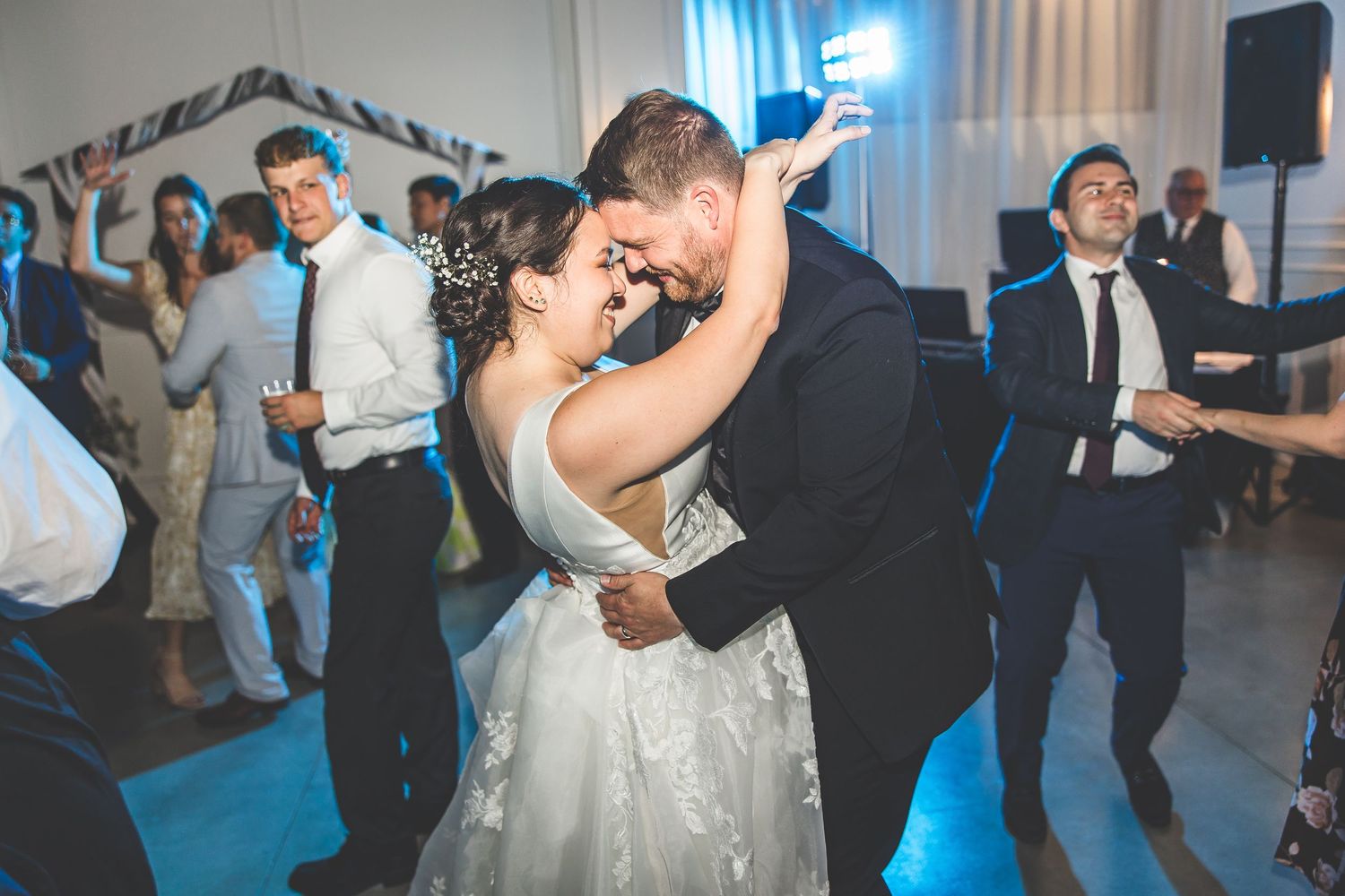 Wedding party dancing enthusiastically under blue mood lighting at an indoor venue.