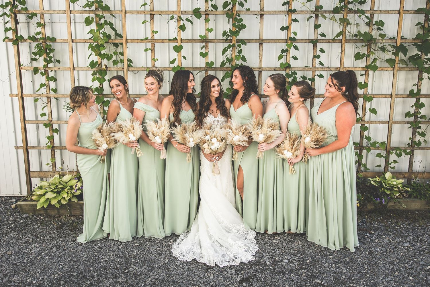 Bridesmaids in mint green dresses stand with bride in white lace gown against lattice wall with vines.