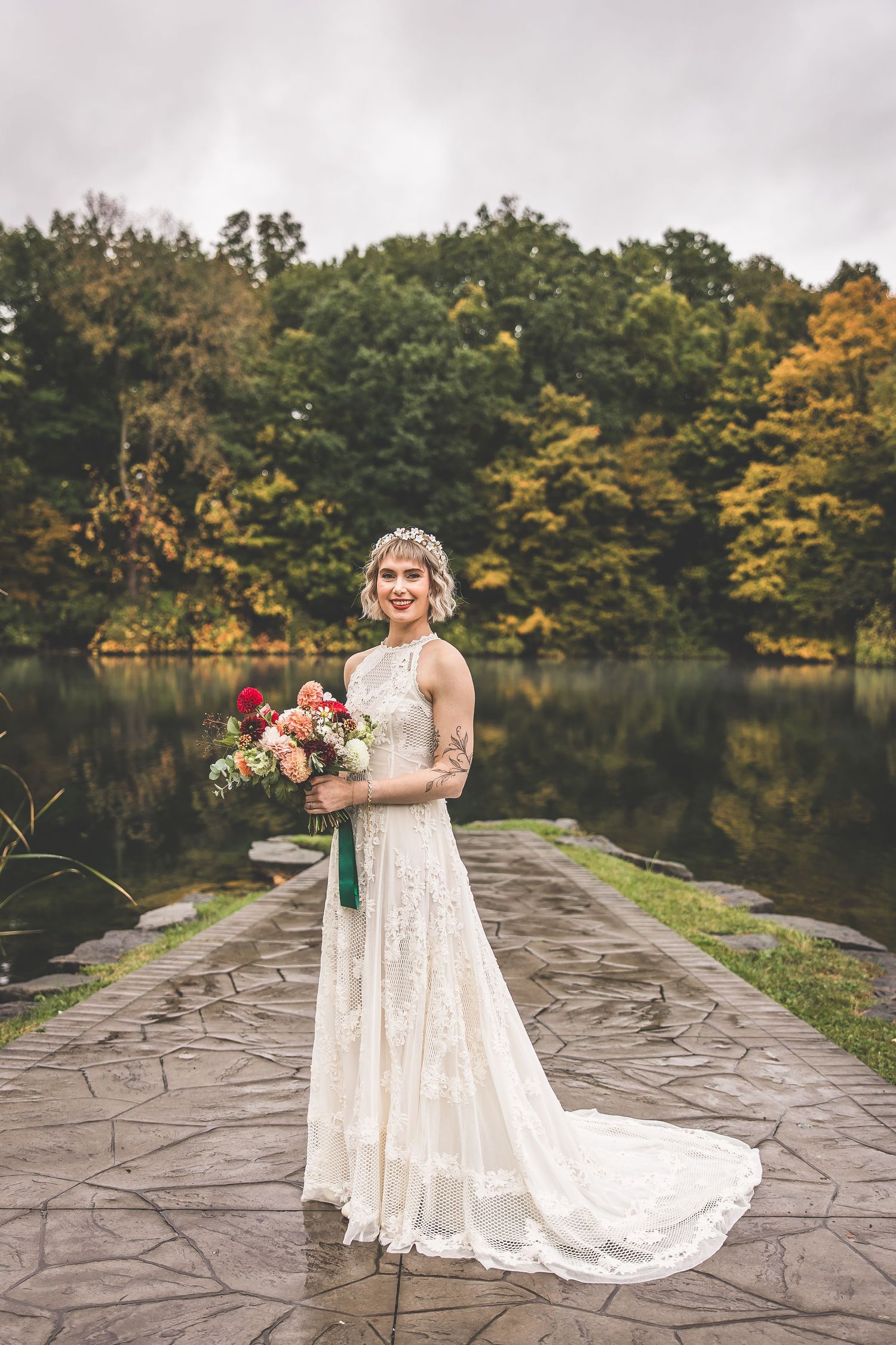 A bride in a lace wedding dress poses on a stone path by a lake surrounded by autumn trees.