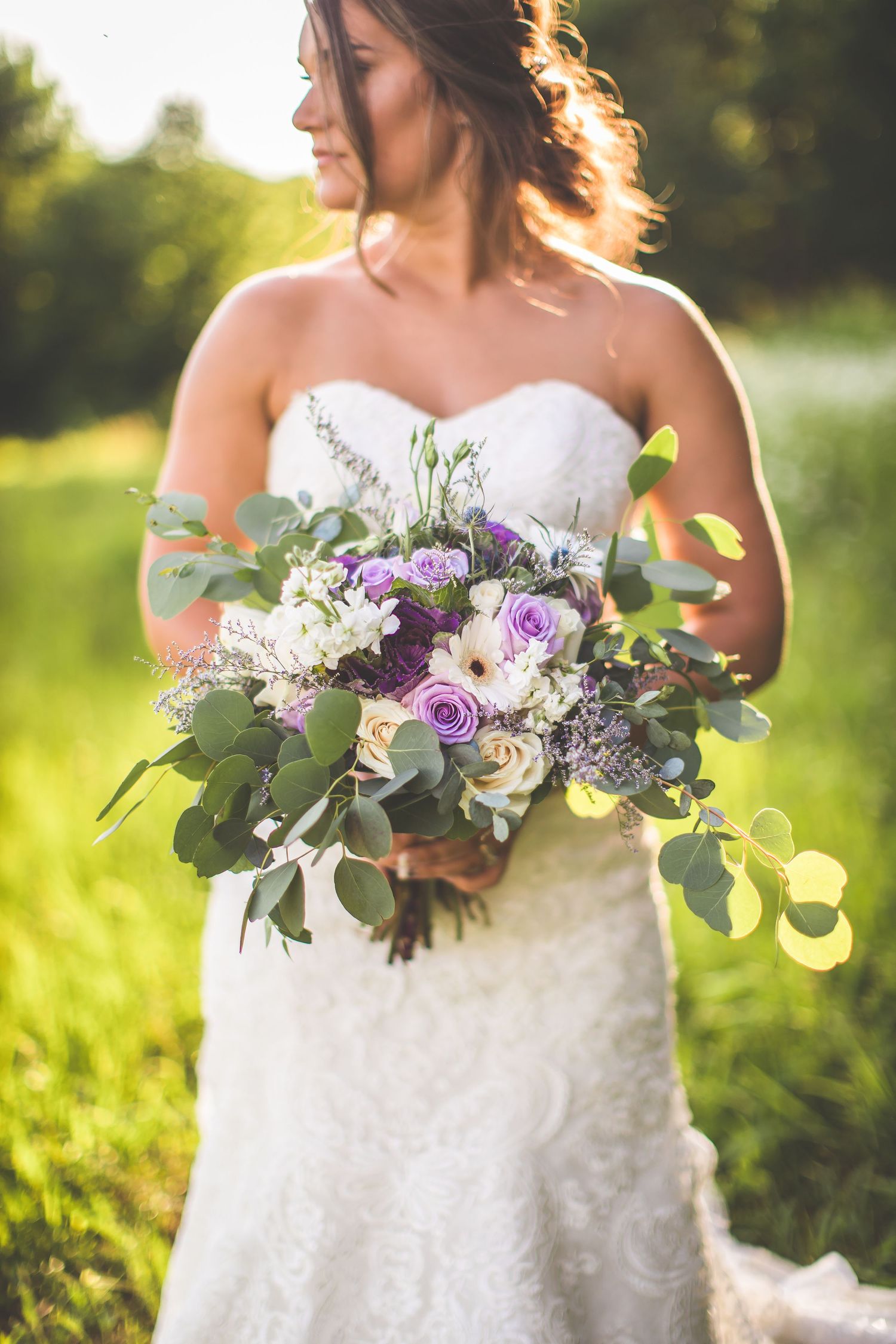 Bridal bouquet with purple and white flowers against sunlit garden background.
