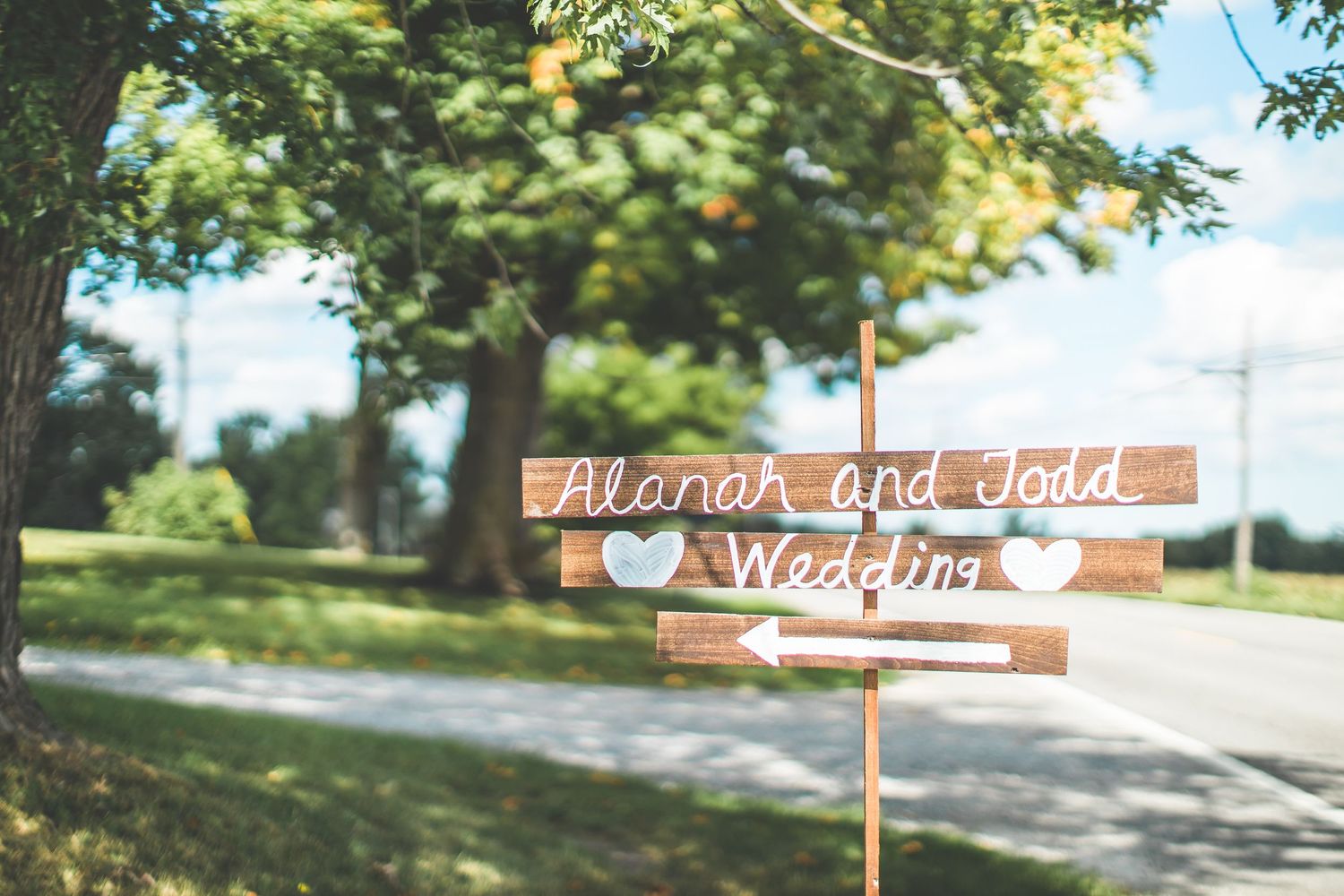 Rustic wooden wedding directional sign with handwritten text against a blurred outdoor background.