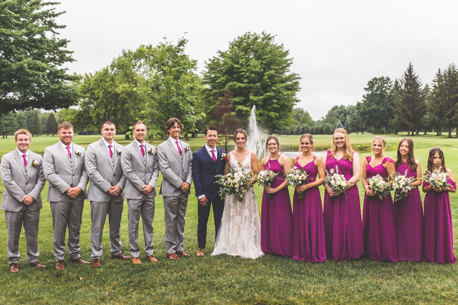 Wedding party group photo with bridesmaids in burgundy dresses and groomsmen in gray suits on a grassy lawn.