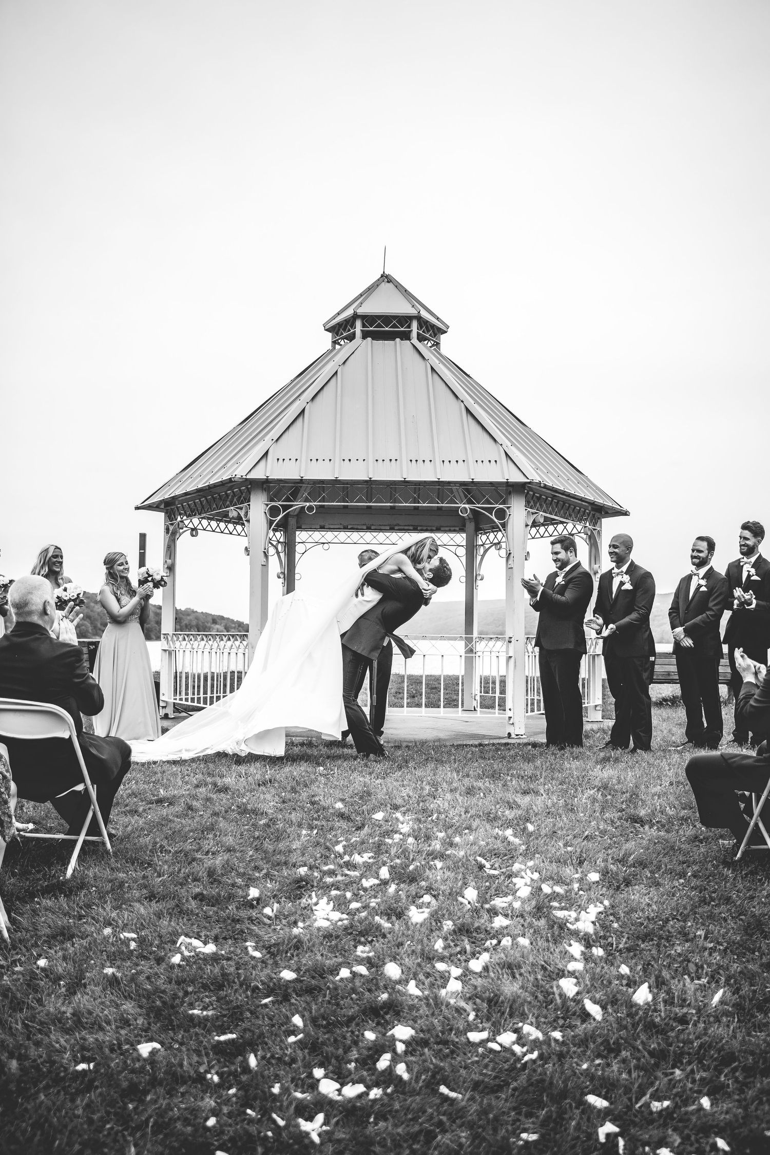 Black and white panoramic view of an outdoor wedding ceremony setup with a gazebo and guests on a grassy hillside.