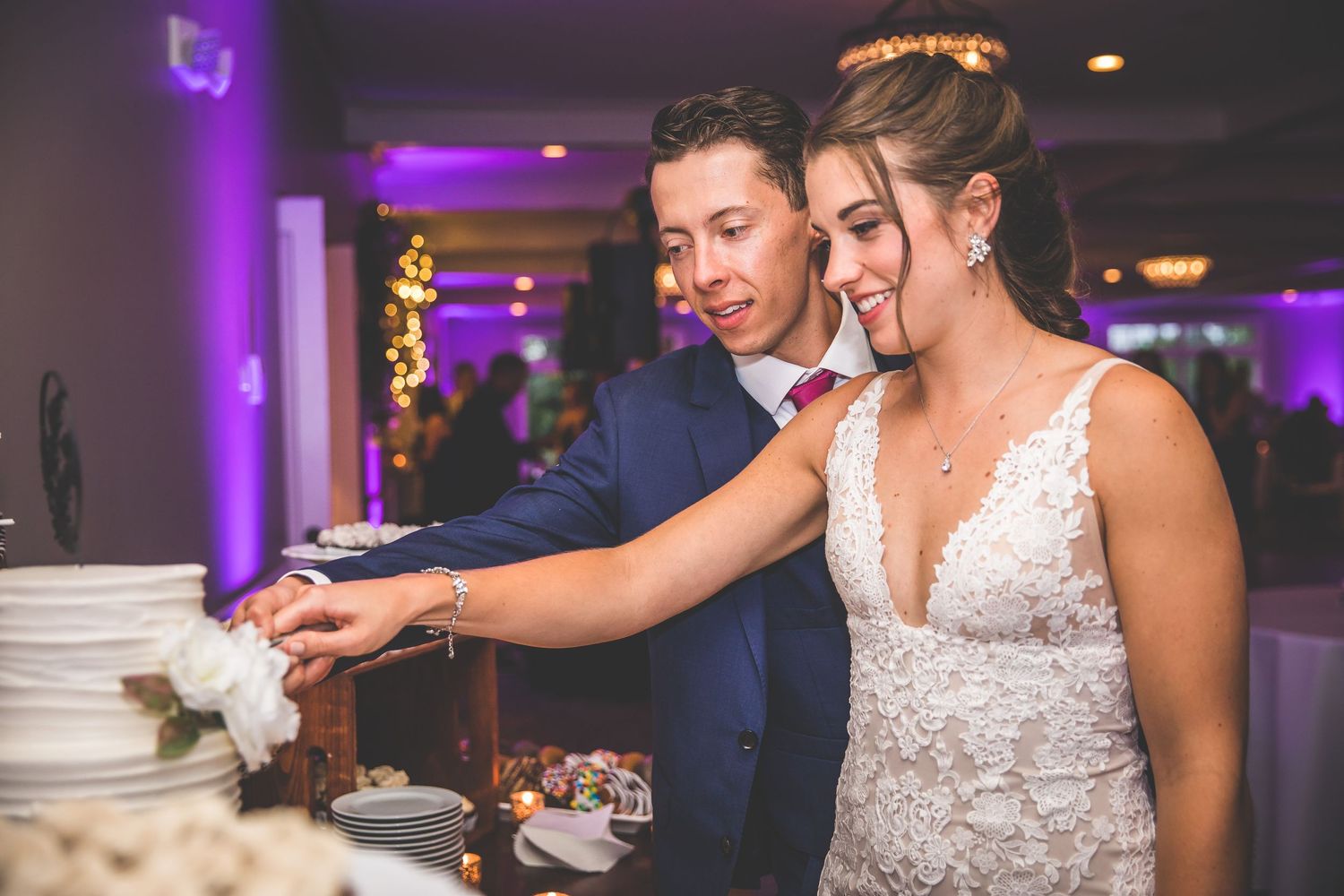 Newlyweds feed each other wedding cake during their reception in a purple-lit ballroom.