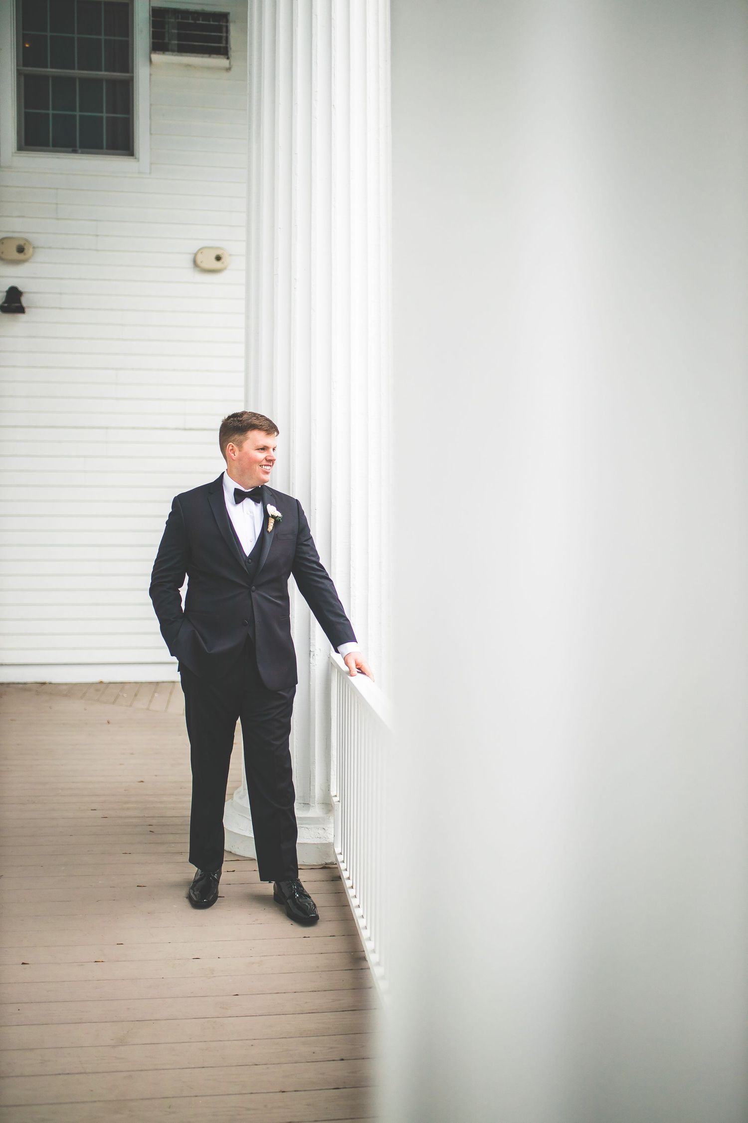 Black and white photo of groom in tuxedo walking on covered porch.