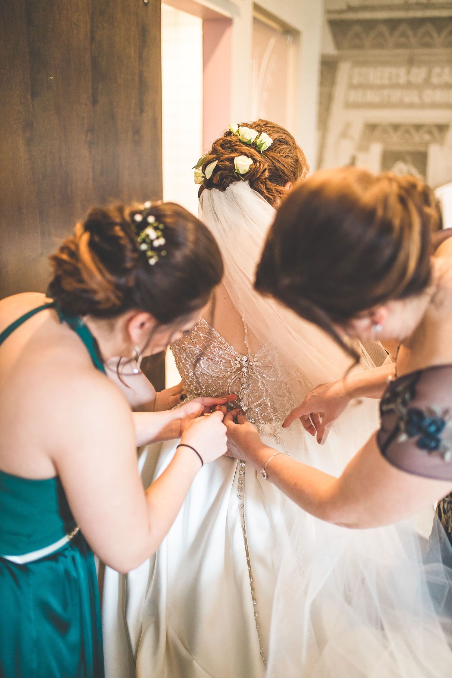Bridesmaids in teal dresses help button up the back of a wedding dress.